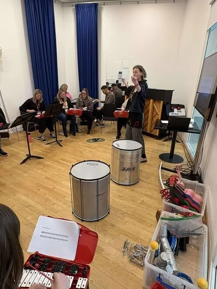 Facilitator standing in front of delegates with arms raised. Delegates sat in semi circle playing glockenspiels.