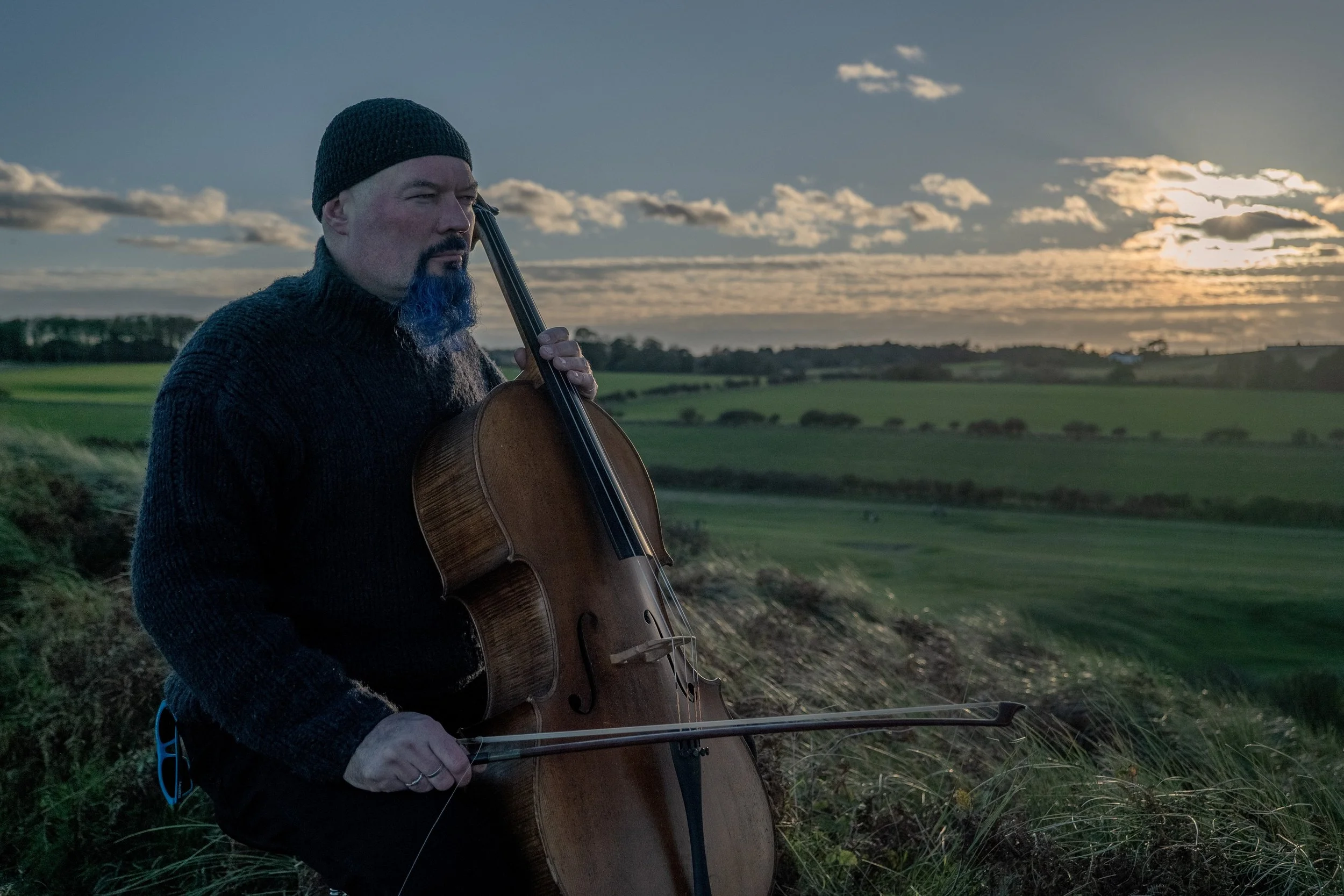 A man in a dark beanie and sweater playing a cello outdoors during sunset over a green landscape.