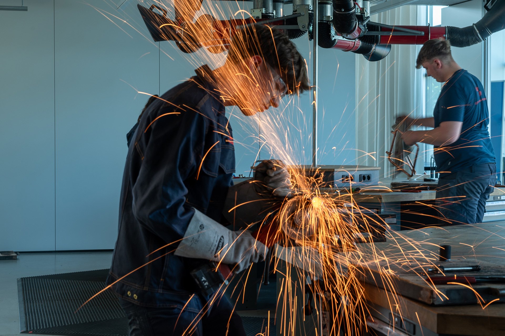 Schweißer bei der Arbeit in einem Werkstatt, während eine Person im Hintergrund eine andere Tätigkeit ausführt.