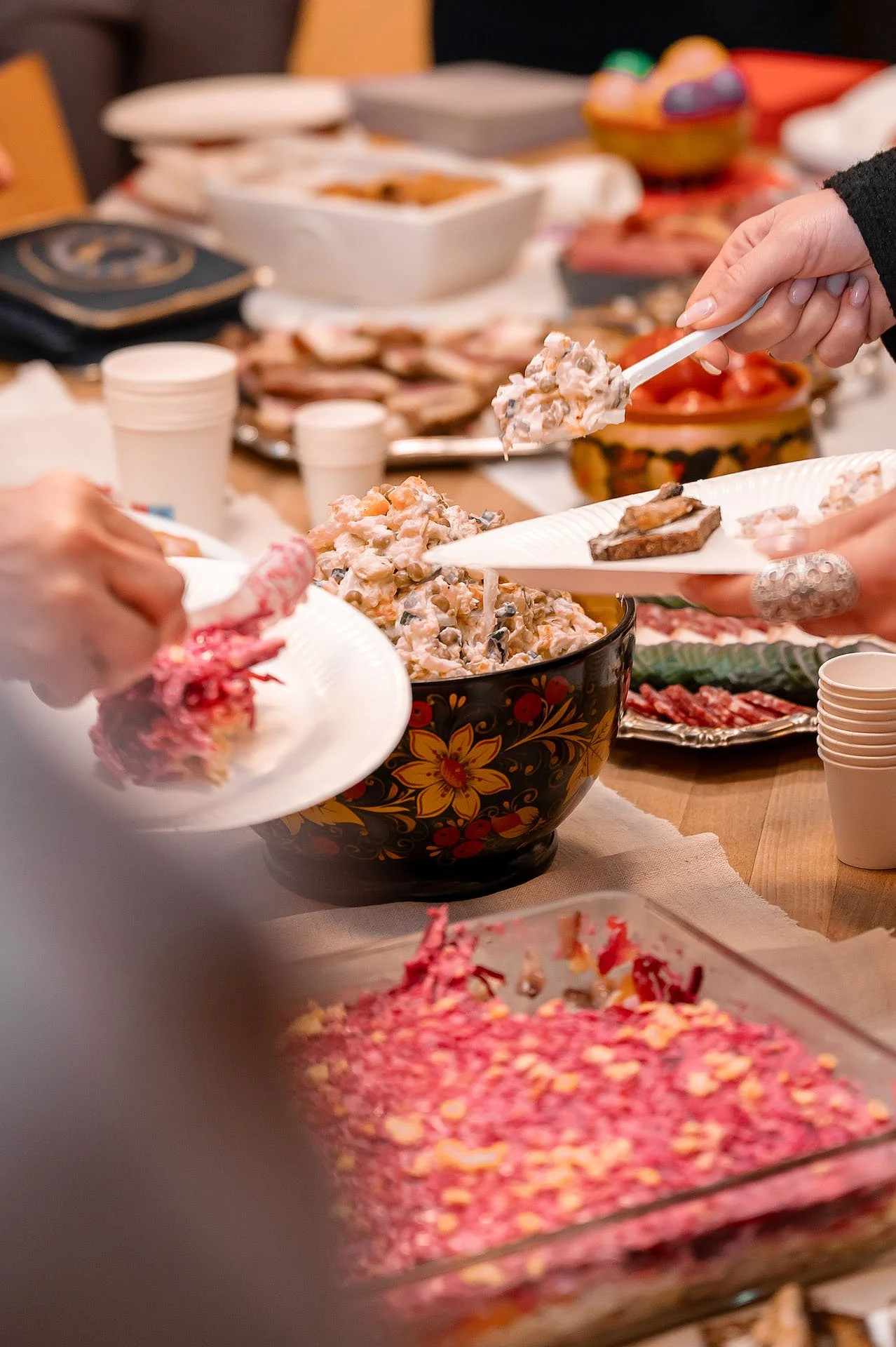 Menschen servieren sich bei einem Gemeinschaftsessen und nehmen Lebensmittel aus einer großen, dekorativen Schüssel mit Salat. Weitere Speisen und Geschirr sind auf einem Tisch sichtbar.
