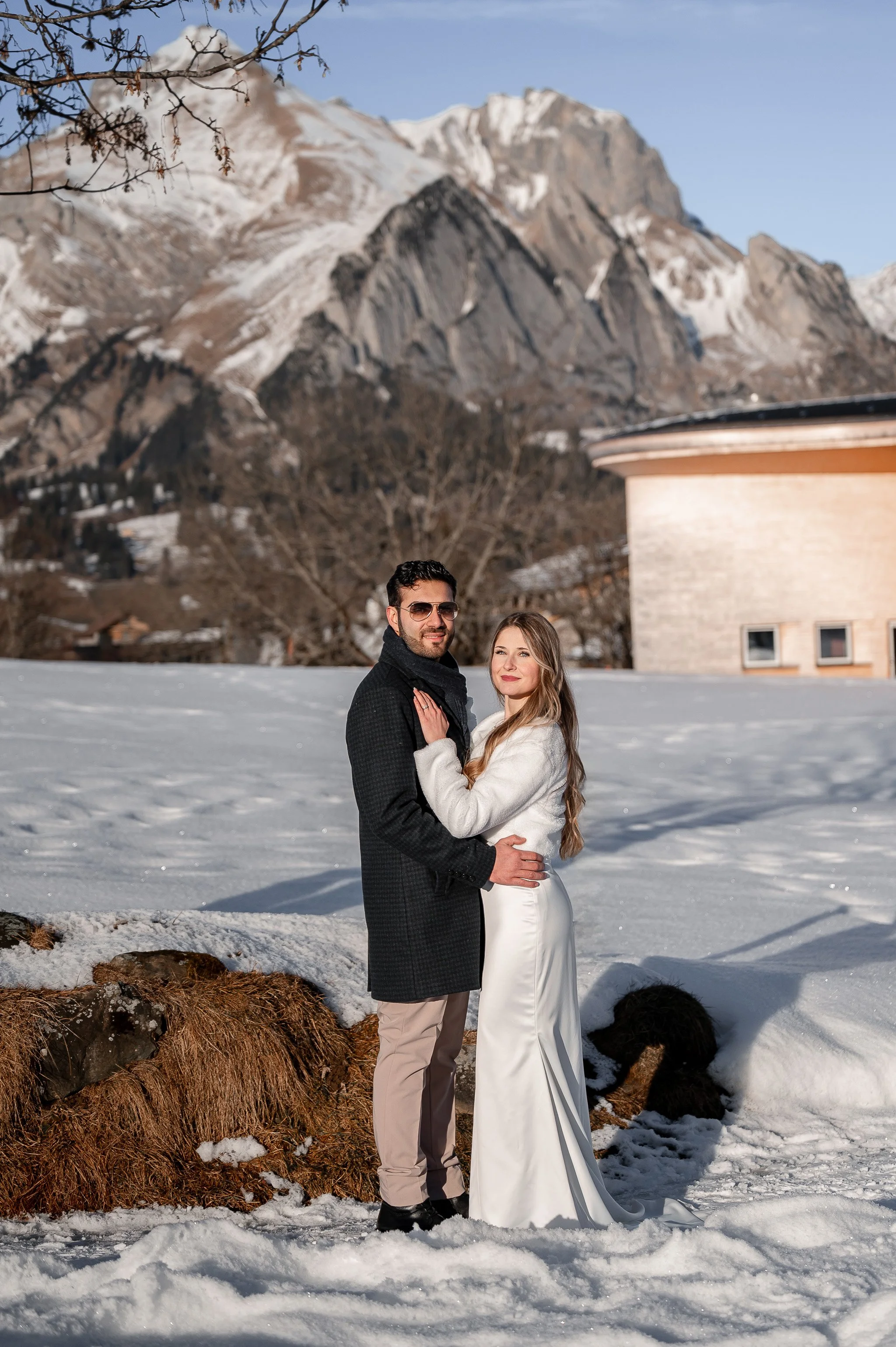 Ein Brautpaar steht im Schnee vor Bergen und Bäumen