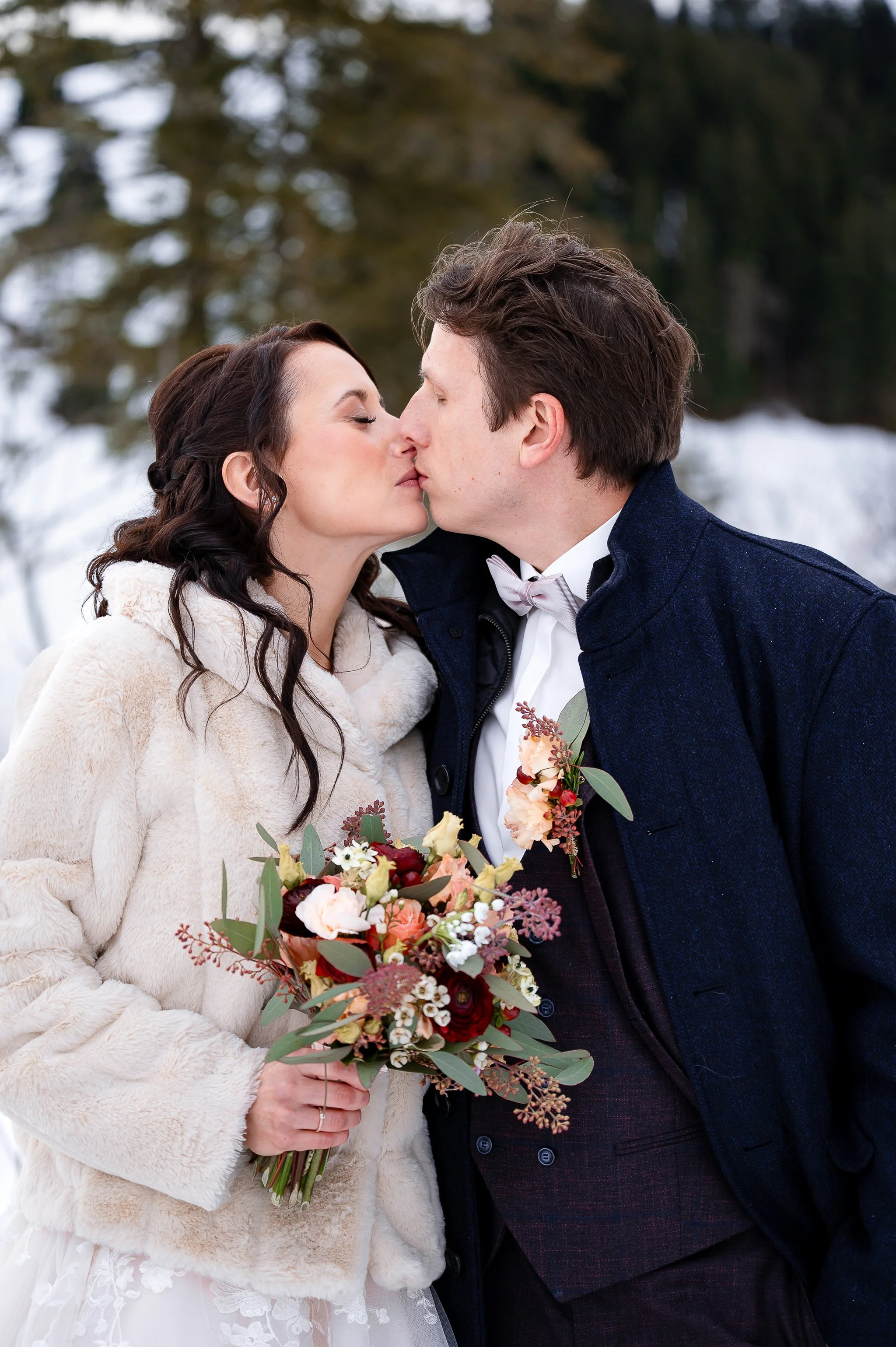 Ein Brautpaar in einer verschneiten Winterlandschaft. Sie küssen sich, die Braut hält einen Blumenstrauß.