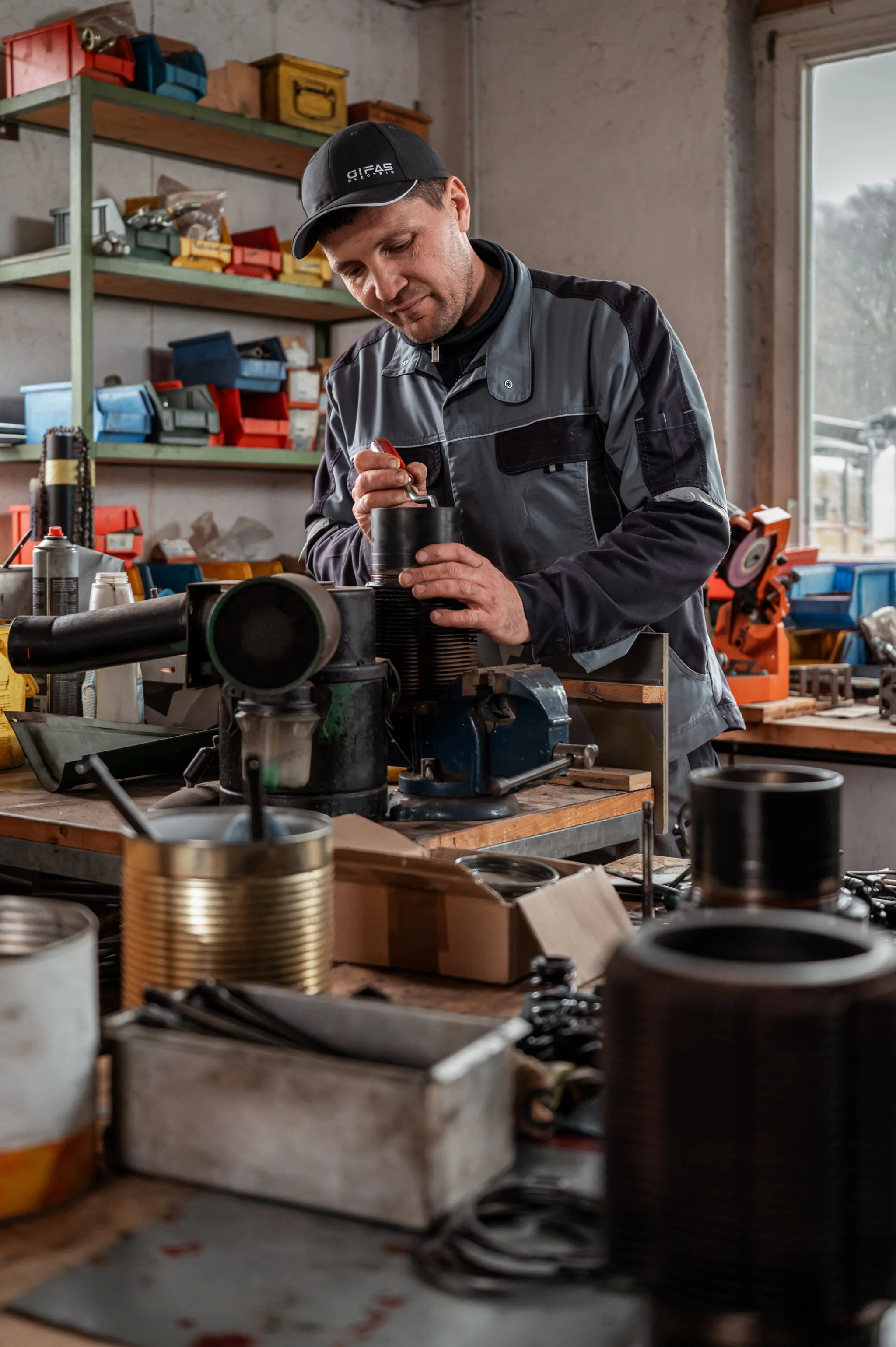 Mann arbeitet in einer Werkstatt, umgeben von Werkzeugen, Werkzeugkisten und Metallteilen.