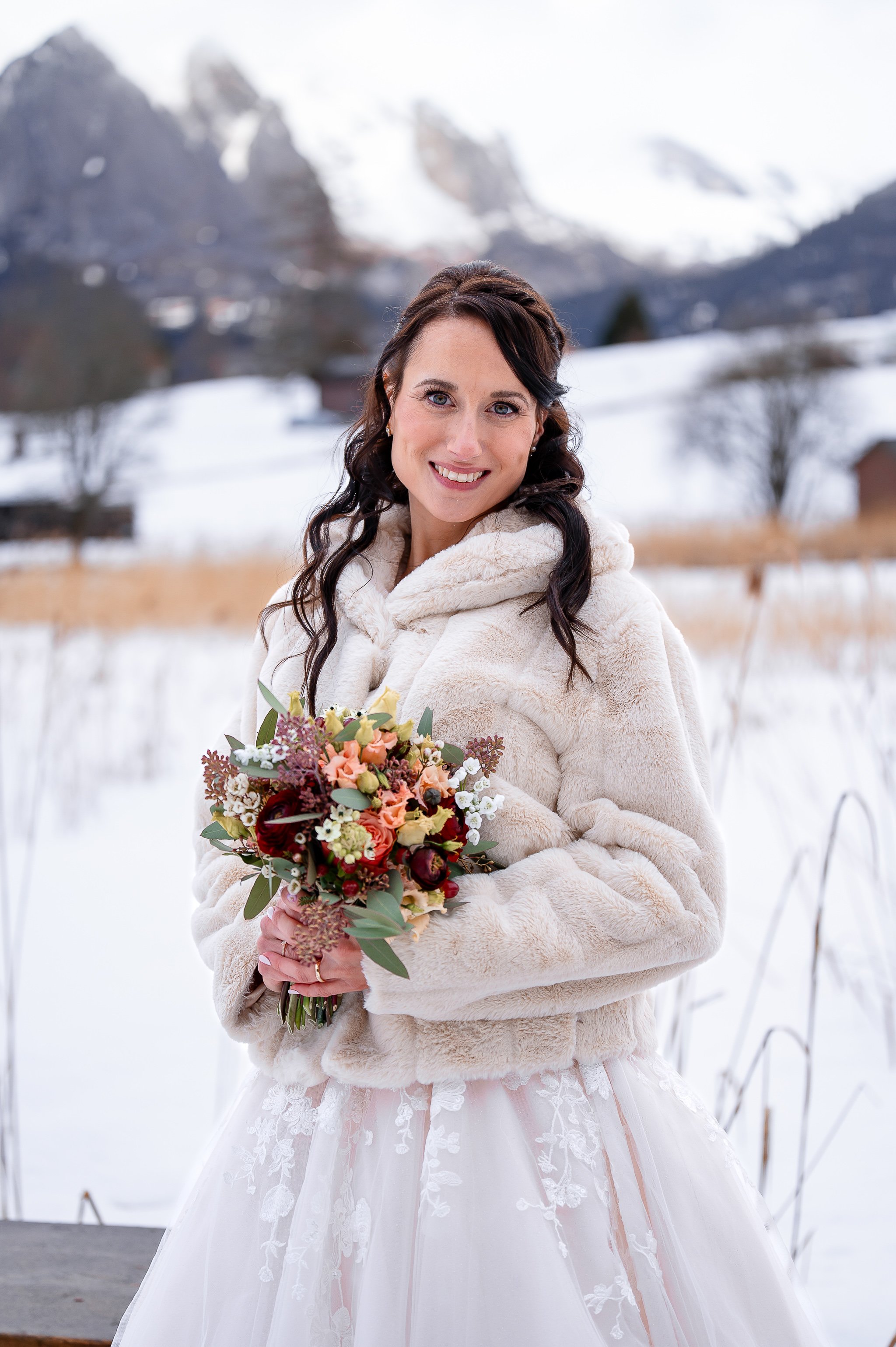 Eine lachende Frau im Brautkleid hält einen Blumenstrauß, mit winterlicher, verschneiter Landschaft im Hintergrund.