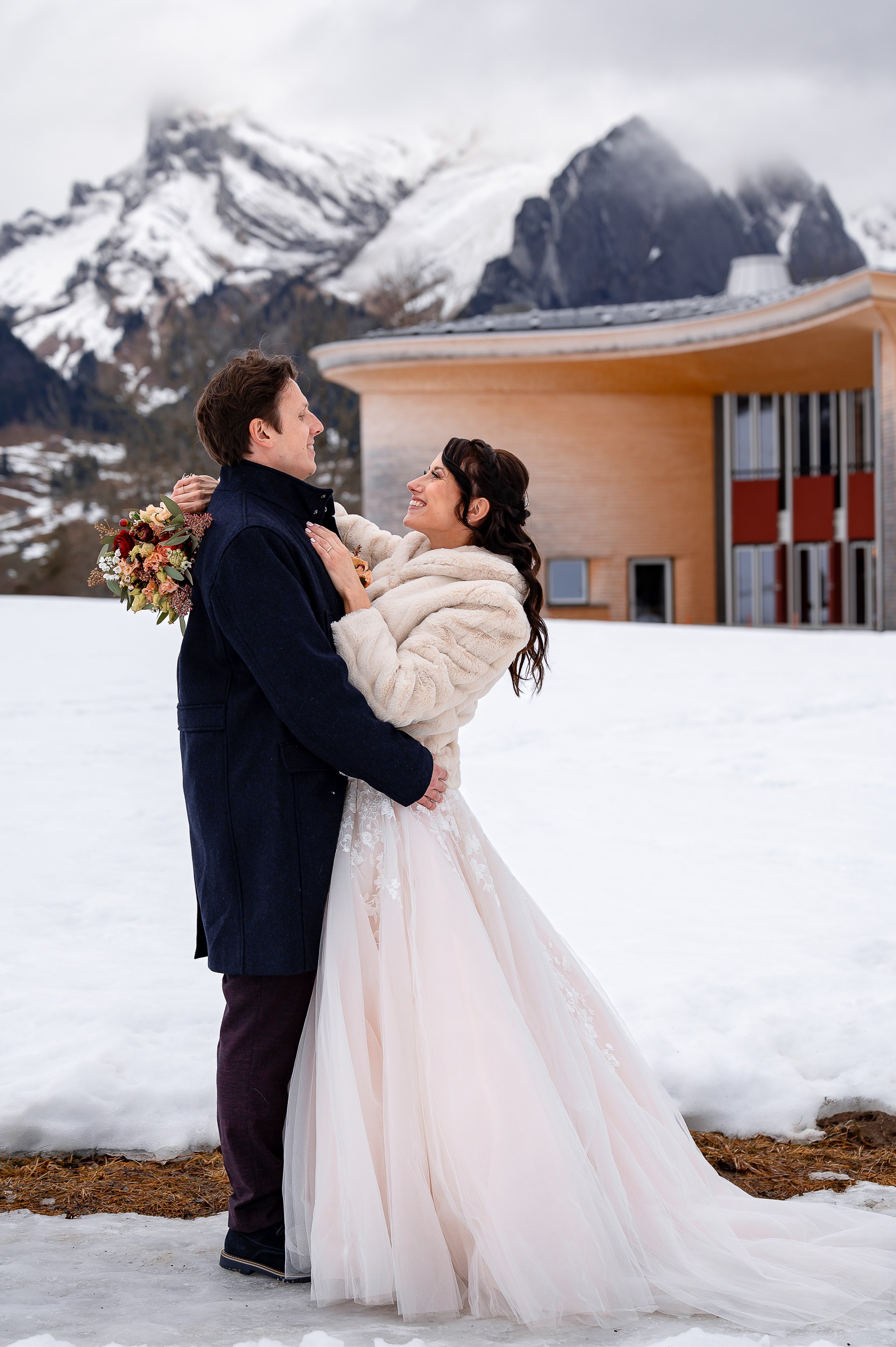 Ein Brautpaar in Winterkleidung tanzt vor einem modernen Haus in schneebedeckter Berglandschaft.