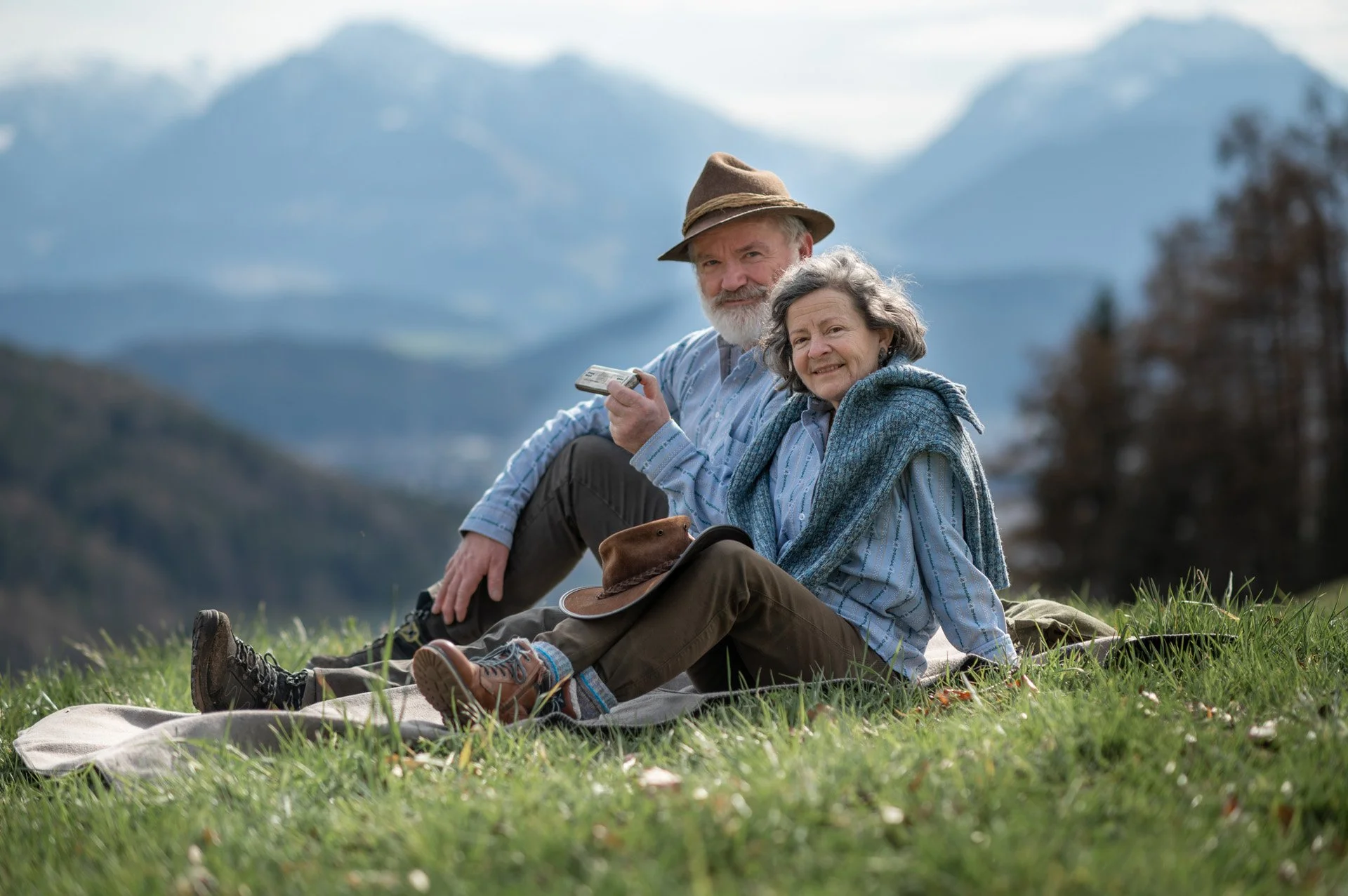 Älteres Paar sitzt entspannt auf einer Wiese mit Blick auf Berge, trägt Naturkleidung, hat Hüte, und genießt die Natur.