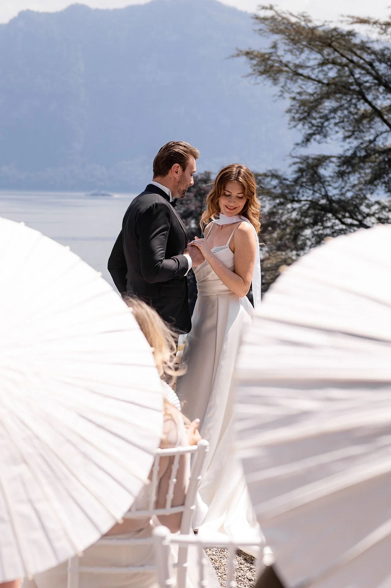 Ein verliebtes Brautpaar bei einer Hochzeit im Freien, umgeben von weißen Sonnenschirm-Strukturen, mit Wasser und Bergen im Hintergrund.