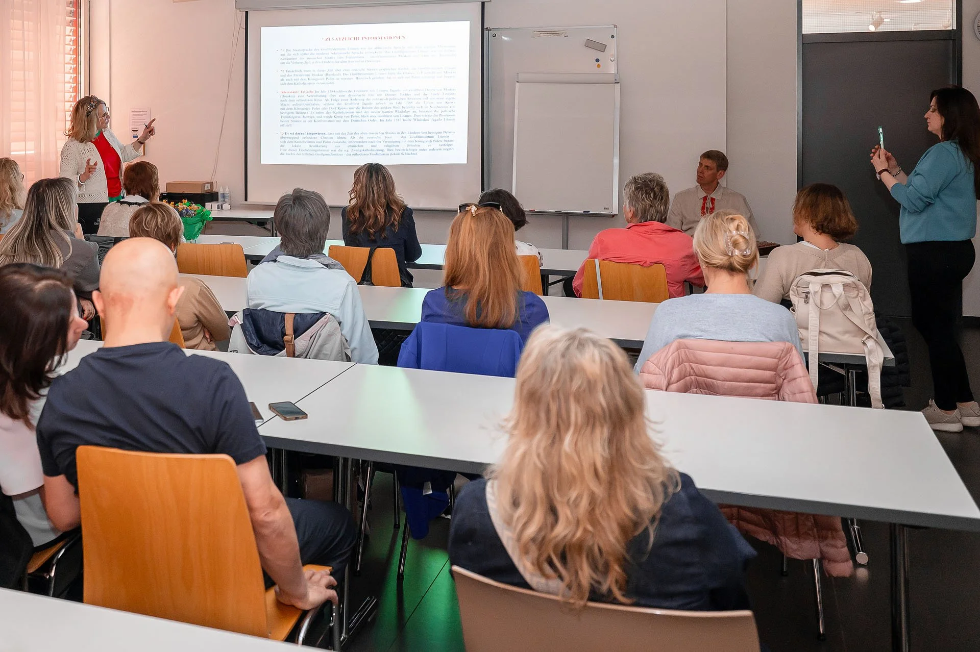 Eine Gruppe von Erwachsenen sitzt in einem Klassenzimmer und hört einer Präsentation zu. Am vorderen Ende steht eine Frau, die mit Gestik spricht, während eine andere Person auf der rechten Seite mit einem Handy den Vortrag aufzeichnet. Es gibt einen