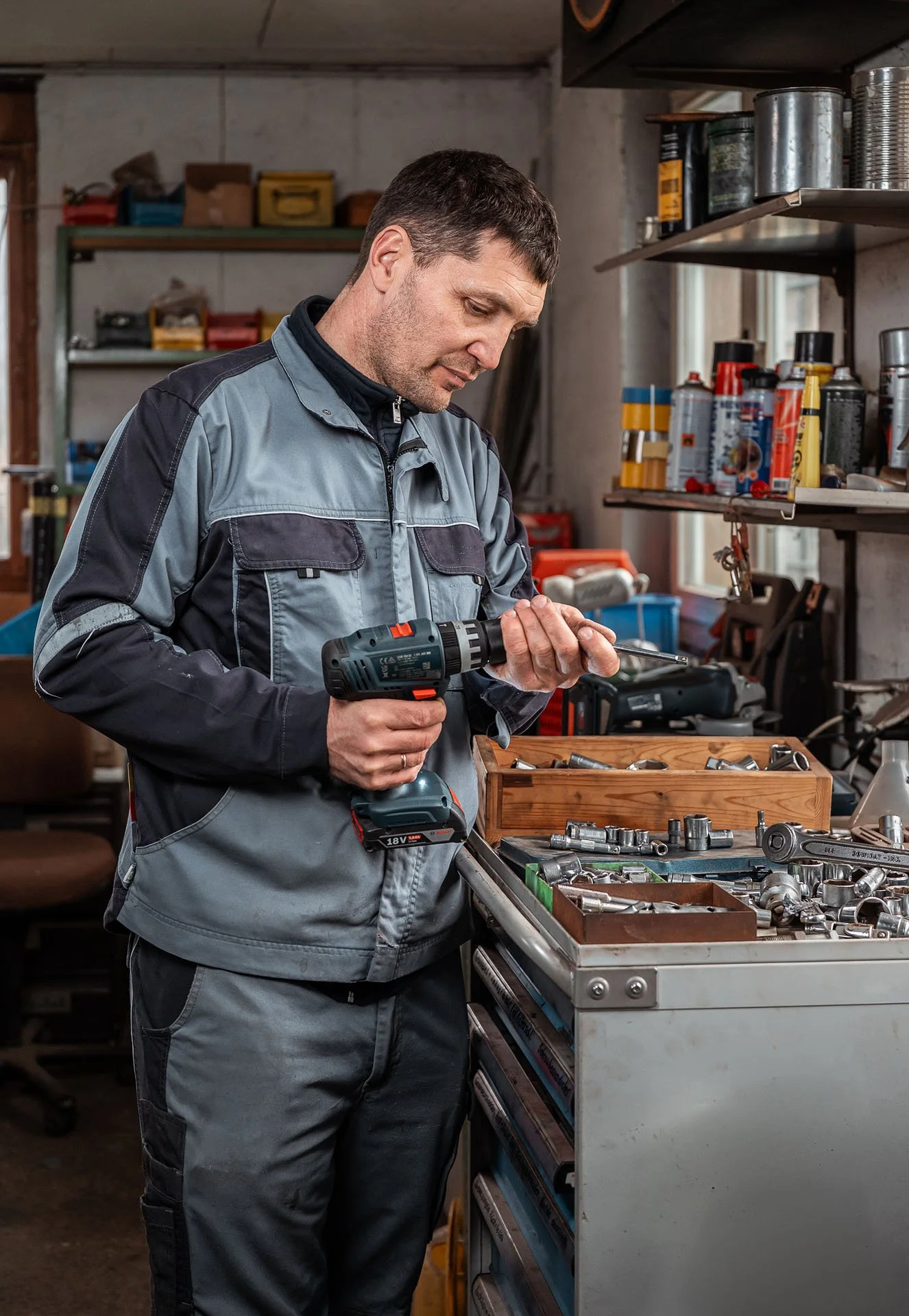 Ein Mann arbeitet mit einem Elektrowerkzeug an einer Werkbank in einer Werkstatt. Es gibt Regale mit verschiedenen Aerosoldosen und Werkzeugen im Hintergrund.