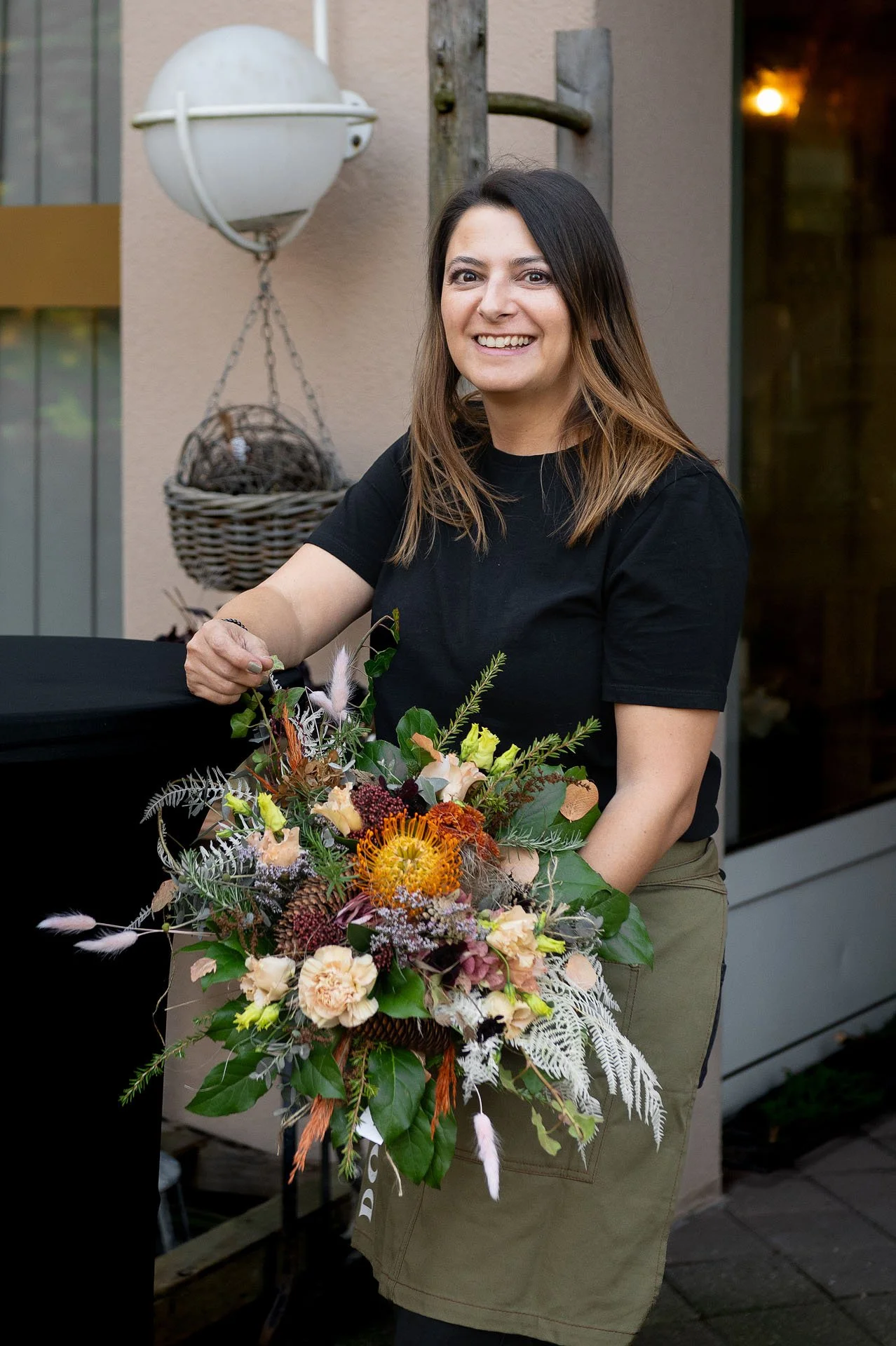 Eine Frau mit braunen Haaren in einem schwarzen T-Shirt und einer grünen Schürze hält einen Blumenstrauß mit bunten Blumen und grünen Blättern. Sie lächelt in die Kamera, im Hintergrund sind ein Fenster, eine Lampe und eine Wand zu sehen.