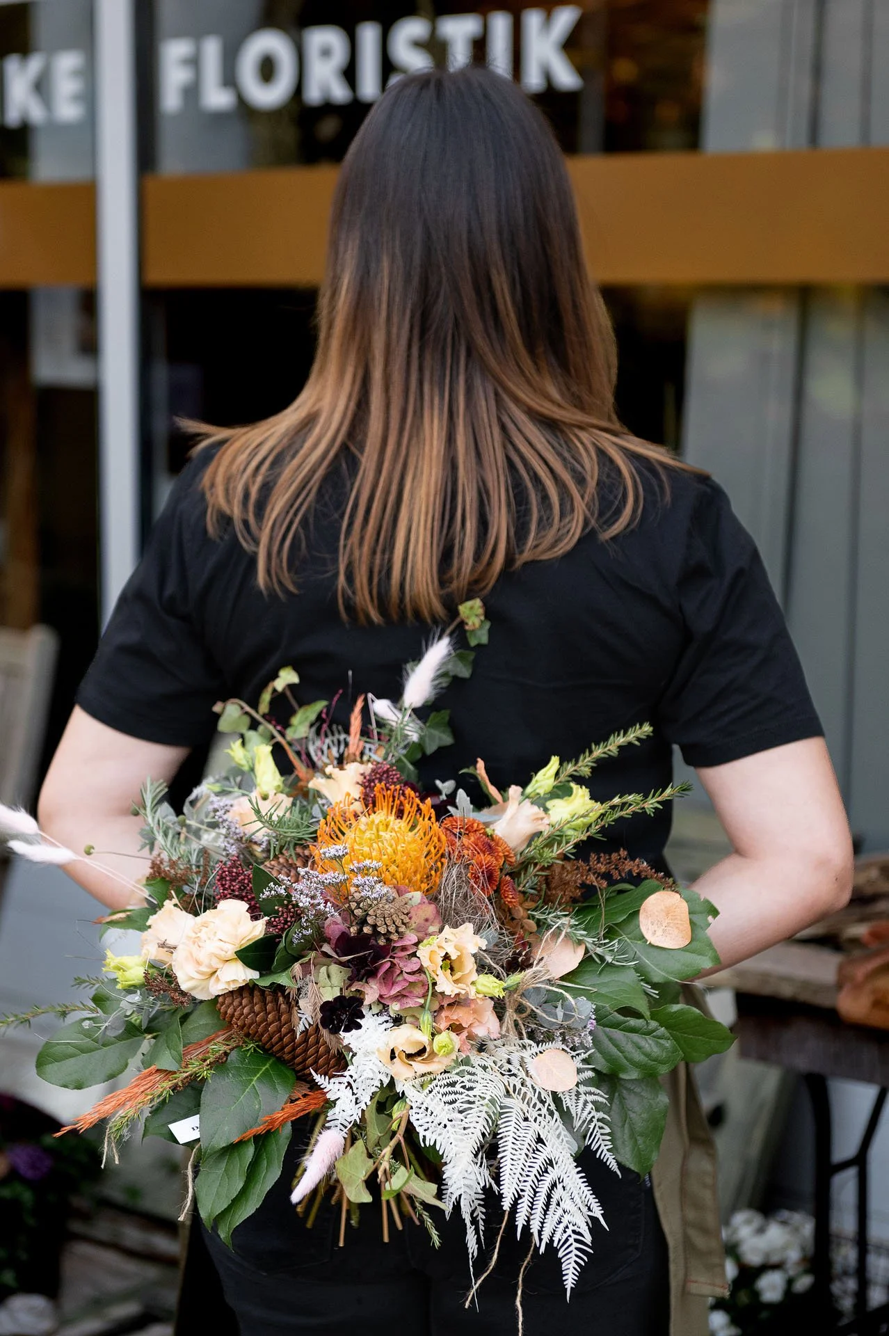 Frau mit mittellang braunem Haar, die einen großen bunten Blumenstrauß trägt, steht vor einem Laden mit der Beschilderung 'Blumistik'.