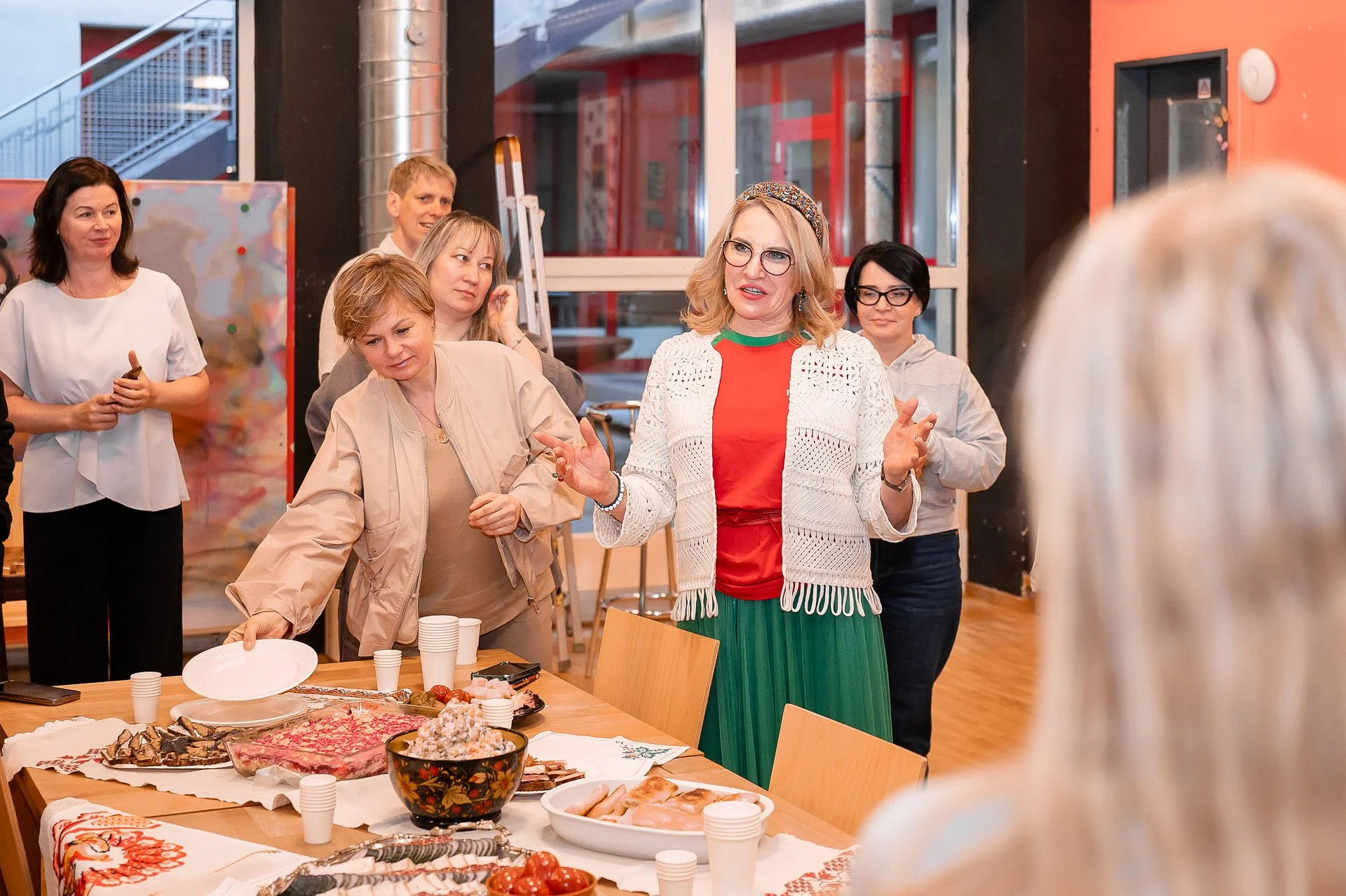 Menschen bei einer Feier mit einem Buffet, das verschiedene Speisen wie Fisch, Salate und Getränke auf einem Tisch präsentiert. Eine Frau in roten und grünen Kleidung spricht, während andere zuschauen.