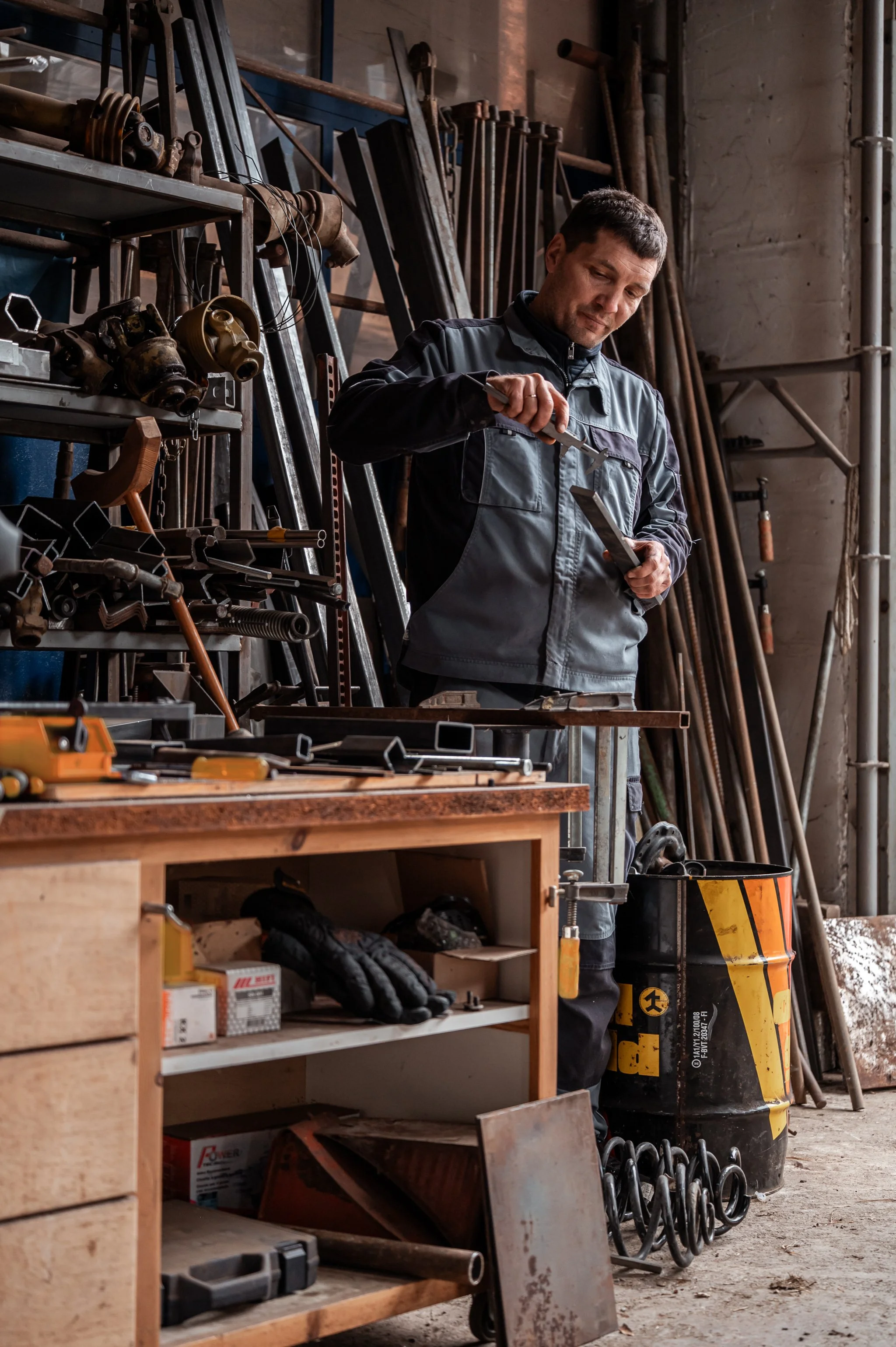 Ein Handwerker in einer Werkstatt, der an einem Metallstück arbeitet, umgeben von verschiedenen Werkzeugen und Werkstücken auf Regal und Arbeitstisch.