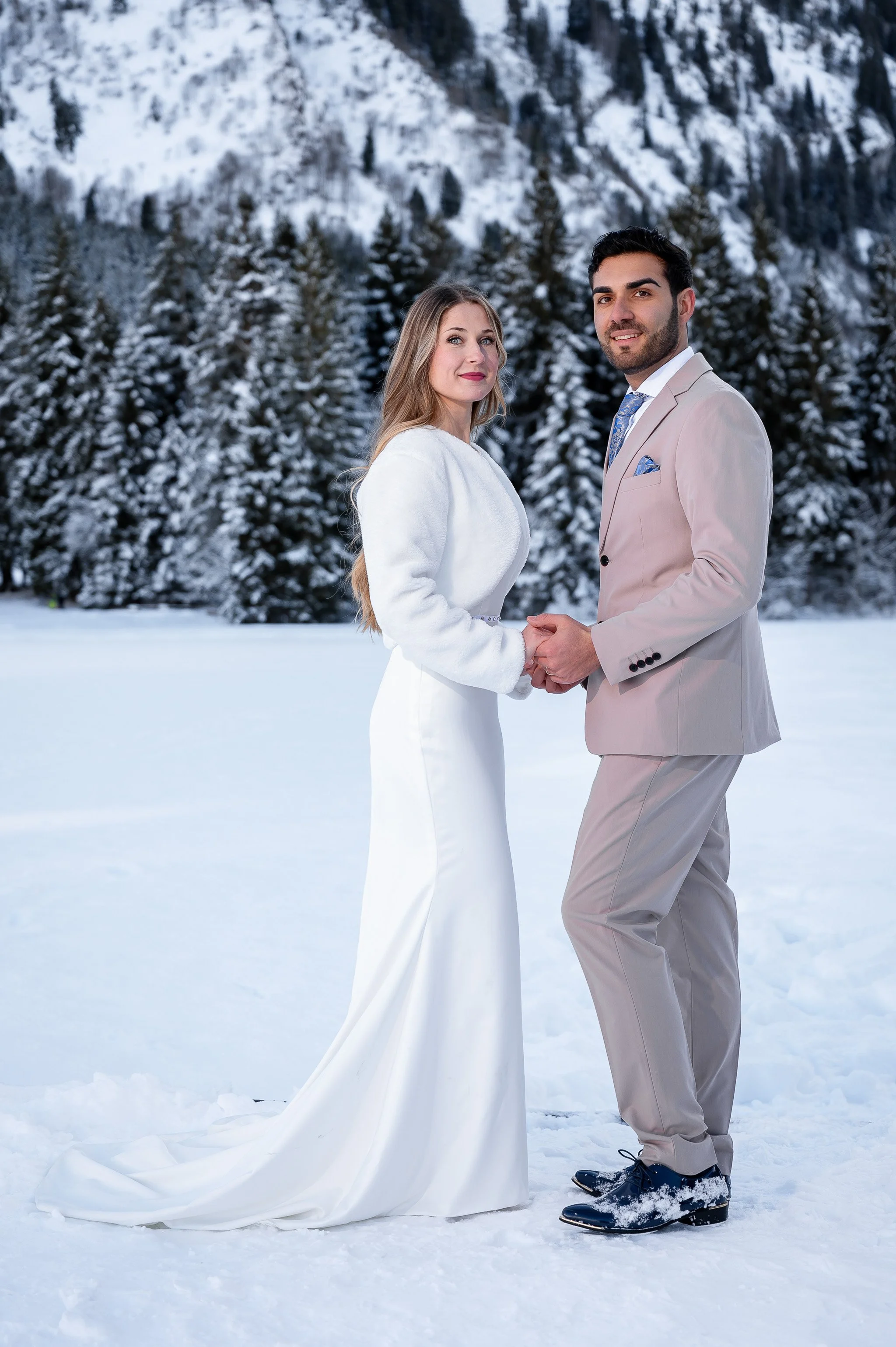 Ein Brautpaar in formeller Kleidung hält sich in einer verschneiten Winterlandschaft an den Händen.