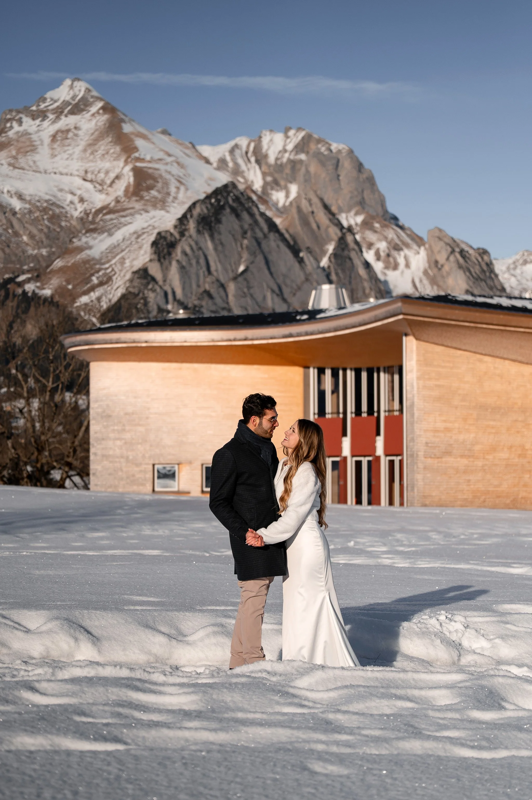 Ein Brautpaar in Winterkleidung, das sich in Schneelandschaft vor einem modernen Gebäude mit Bergen im Hintergrund küsst.