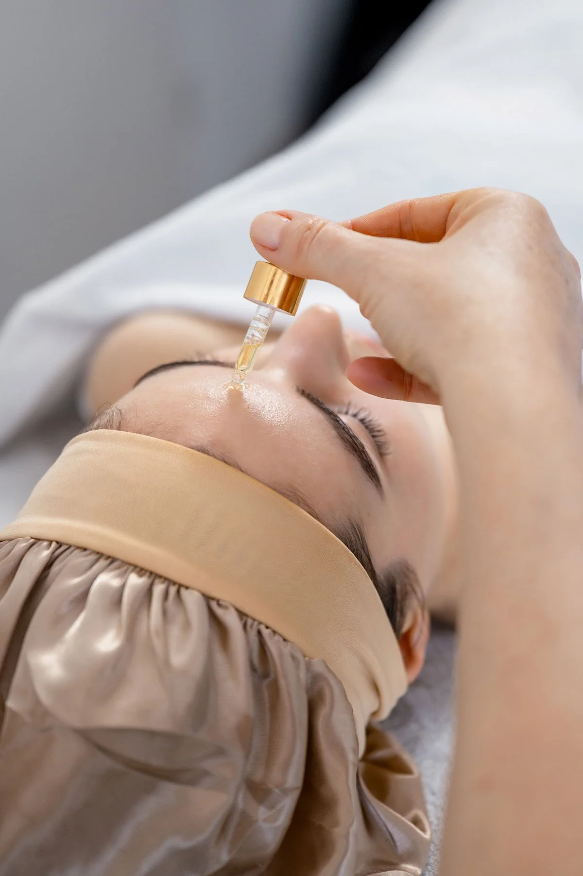Eine Person erhält eine Gesichtspflegebehandlung mit einem Tropfen Serum, während sie mit geschlossenen Augen entspannt auf einer Behandlungsliege liegt, auf einem Kopfband und einem weißen Tuch.