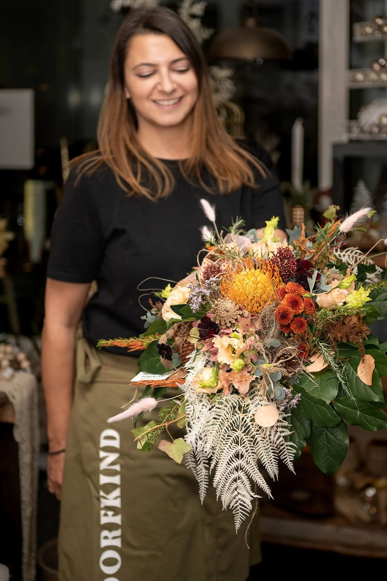 Frau hält einen großen Blumenstrauß mit vielfältigen Blumen und Blättern. Im Hintergrund ein dunkler Raum mit Dekorationen.
