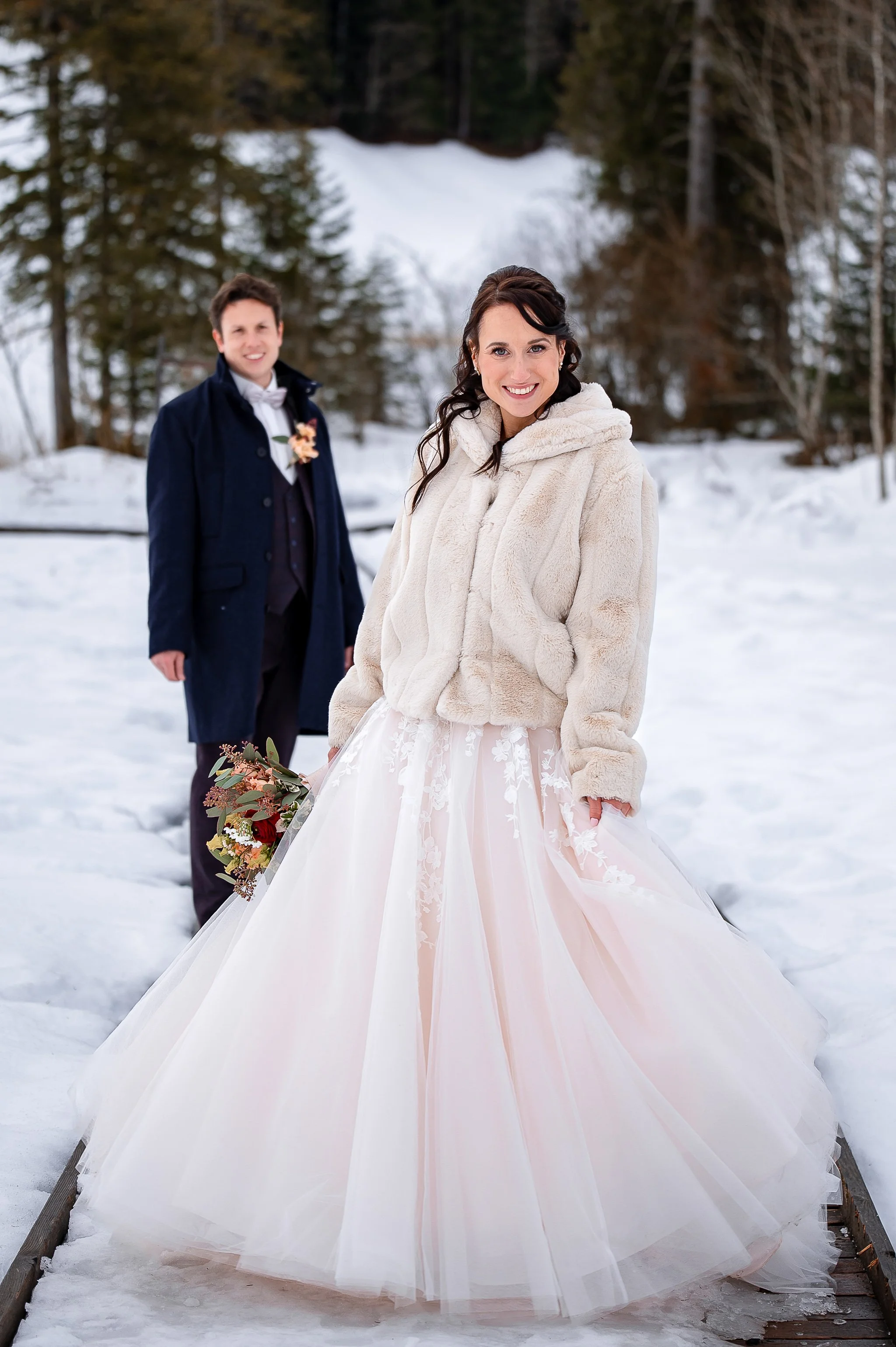 Ein brautpaar in Winterkleidung vor verschneiter Waldkulisse, die Braut trägt ein langes weißes Brautkleid und hält einen Blumenstrauß, während der Bräutigam einen dunklen Anzug trägt.