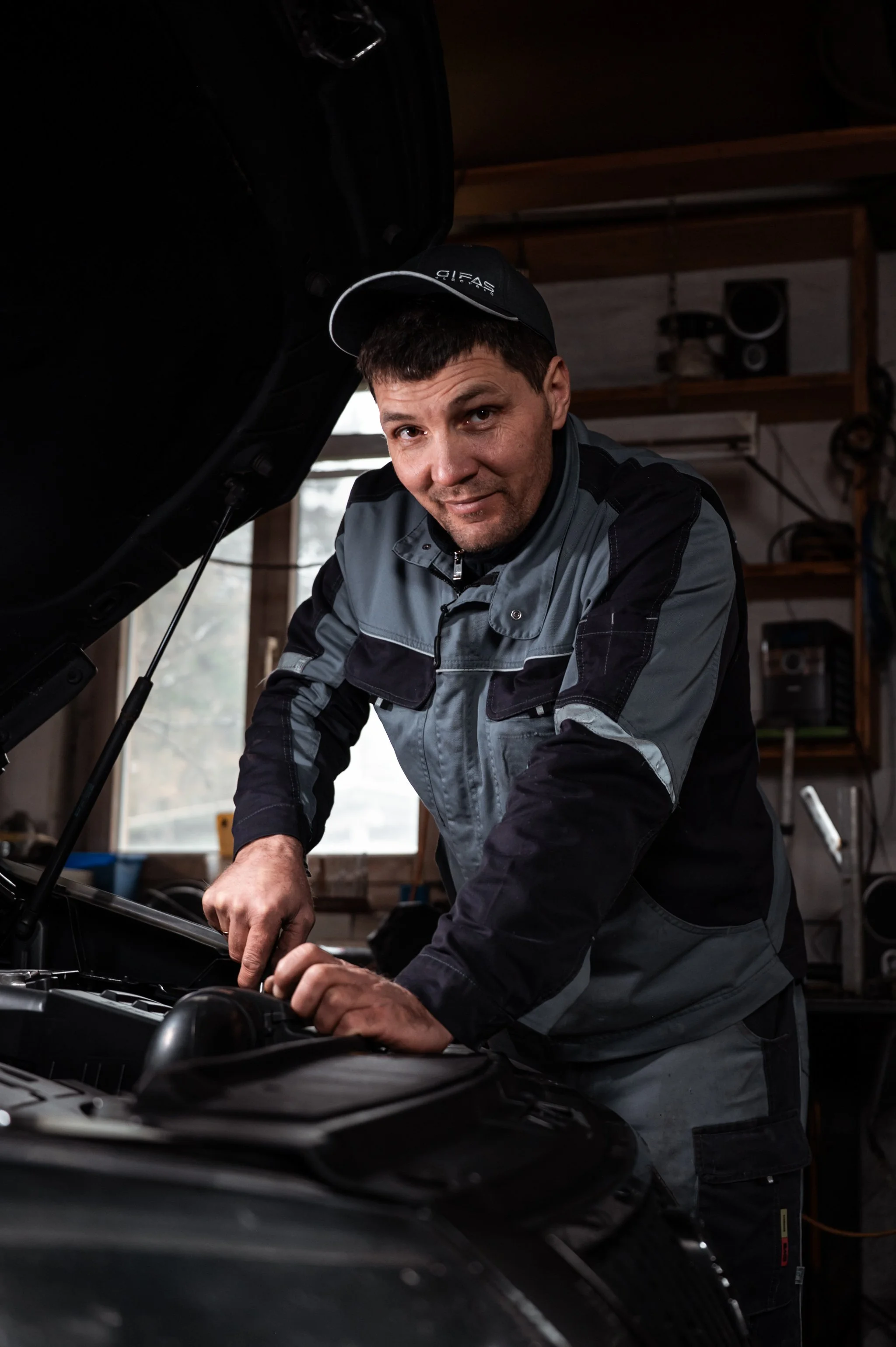 Mann repariert Auto im Garage, arbeitet am Motor unter geöffnetem Kofferraum.