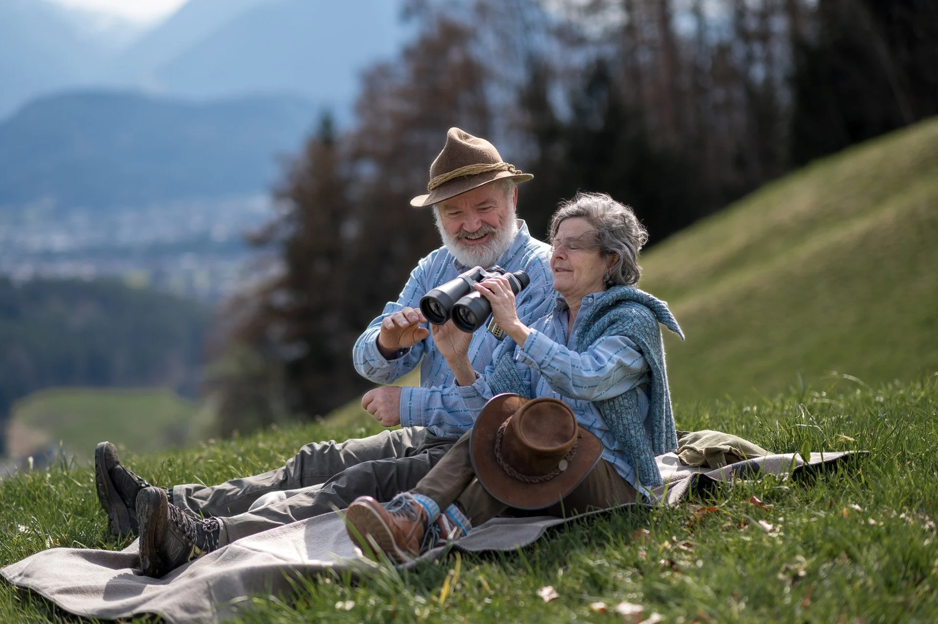 Ein älteres Paar sitzt gemeinsam auf einer Wiese im Freien, schaut durch ein Fernglas und lächelt. Es ist ein sonniger Tag, im Hintergrund sind Berge und Bäume sichtbar.
