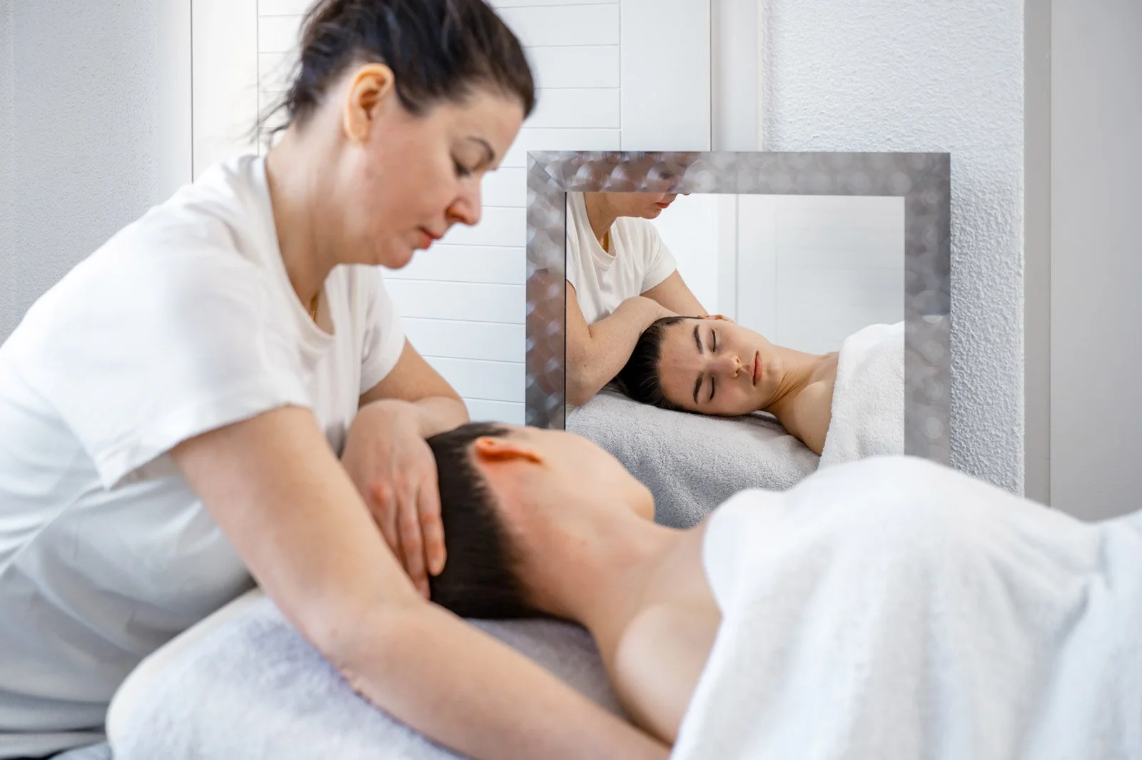 Masseur gibt einer Frau eine Nackenmassage in einem Spa oder Wellnesszentrum, während sie entspannt auf einer Liege liegt und im Spiegel neben ihr zu sehen ist.