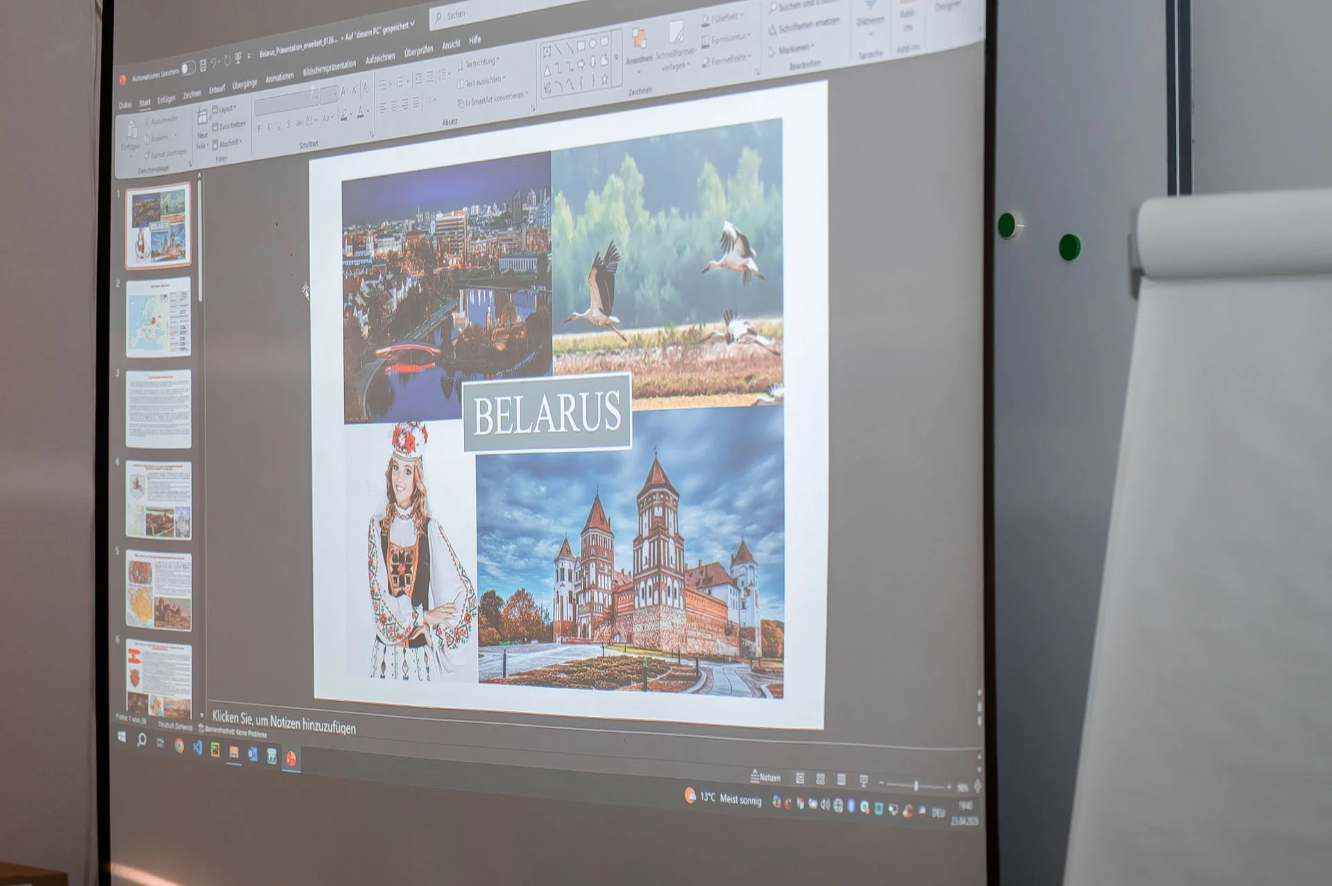 Ein Computerbildschirm zeigt eine Präsentation über Belarus mit Fotos der Stadt, Natur, Wildvögeln, einer Frau in traditioneller Kleidung und einer historischen Kirche.