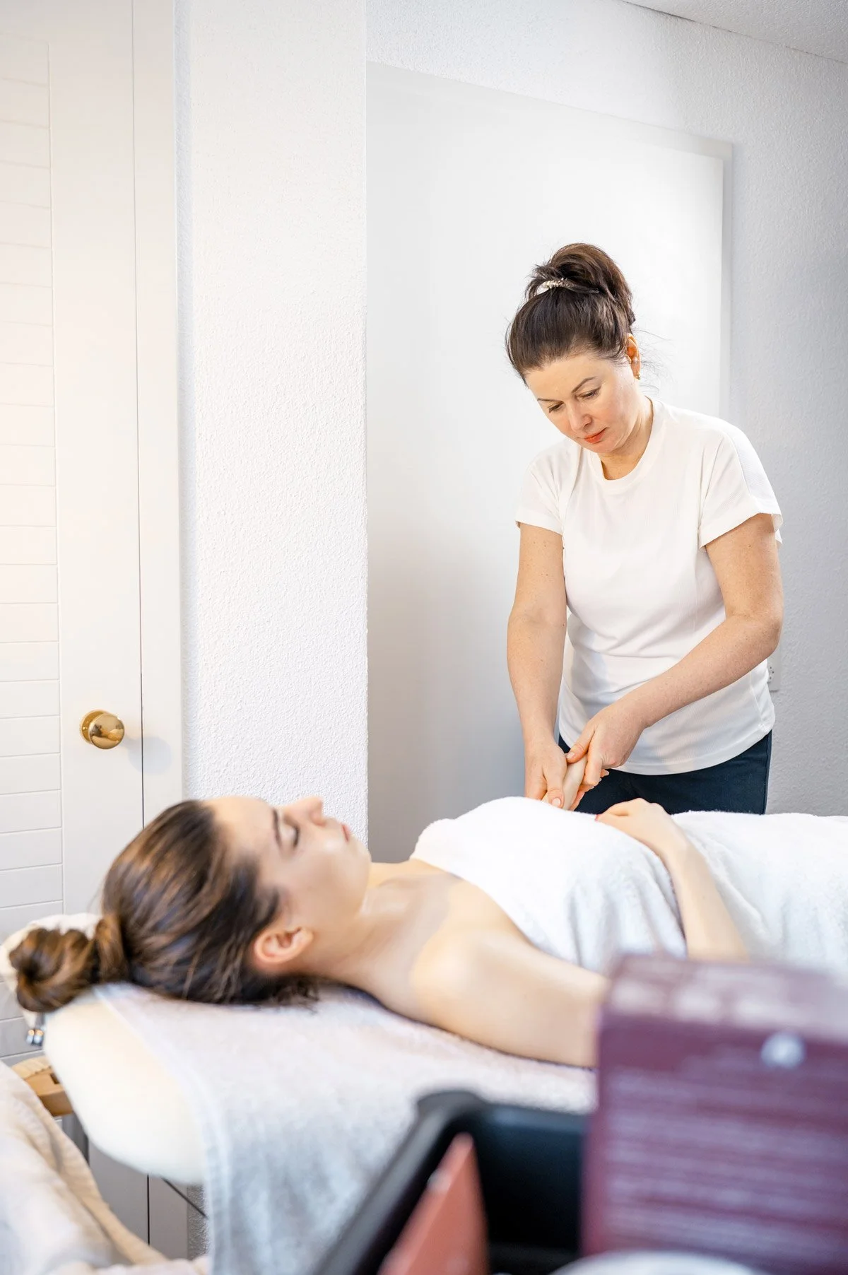 Eine Frau liegt entspannt auf einer Behandlungsliege, während ein Therapeut eine Handmassage an ihrer Hand durchführt in einem Wellness- oder Spa-Bereich.