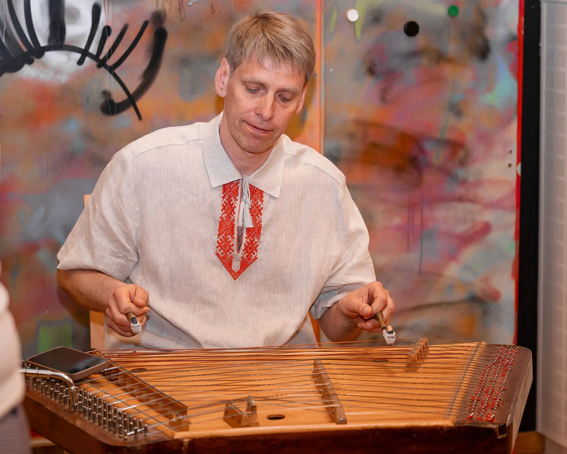 Ein Mann in traditioneller ukrainischer Tracht spielt auf einer Cimbalom, einem käsigen Instrument, in einem Raum mit abstrakter Wandkunst im Hintergrund.