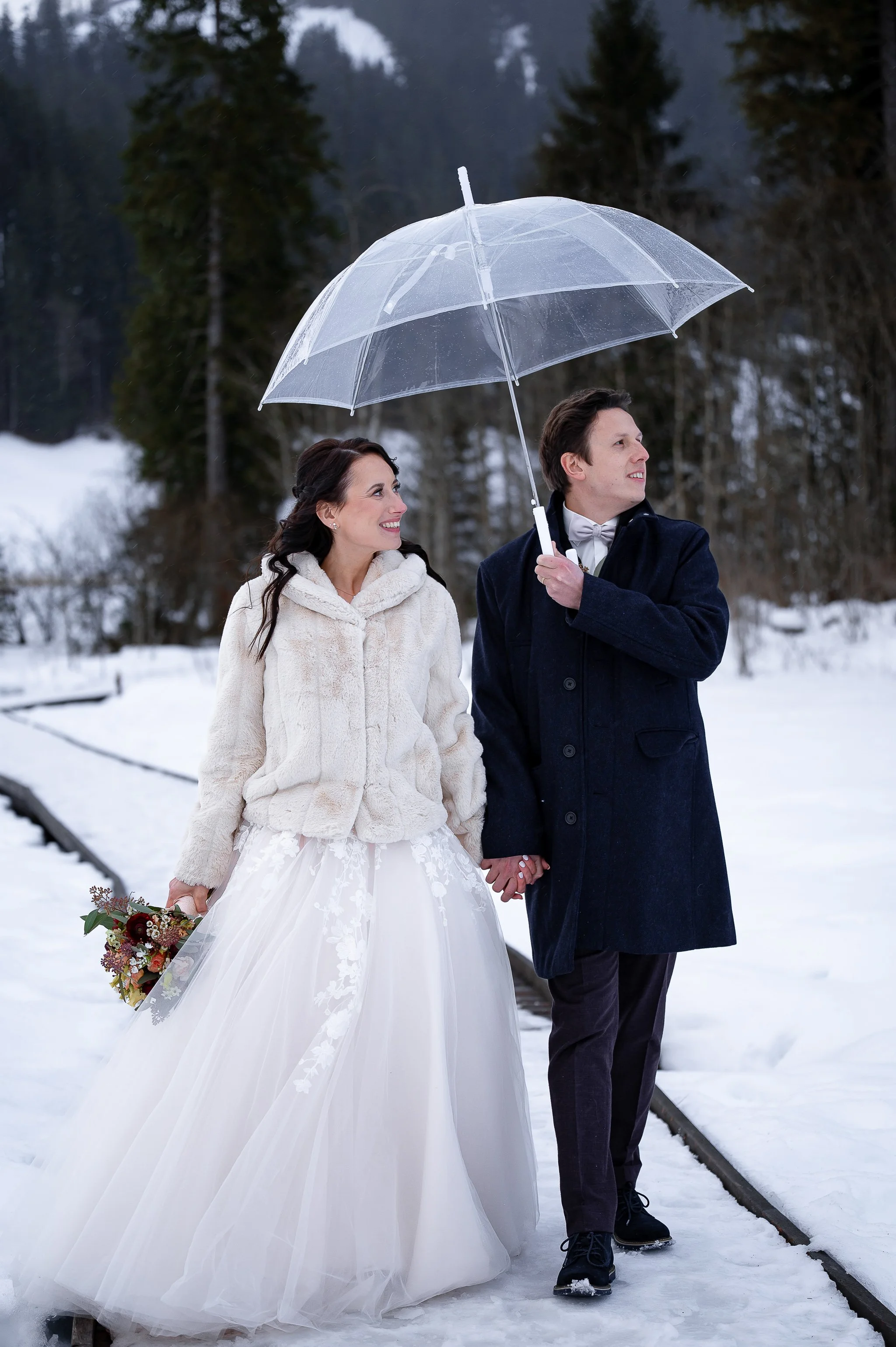 Ein Brautpaar spaziert im Schnee unter einem transparenten Regenschirm, die Frau trägt ein weißes Brautkleid und eine Pelzjacke, der Mann trägt einen dunklen Mantel.