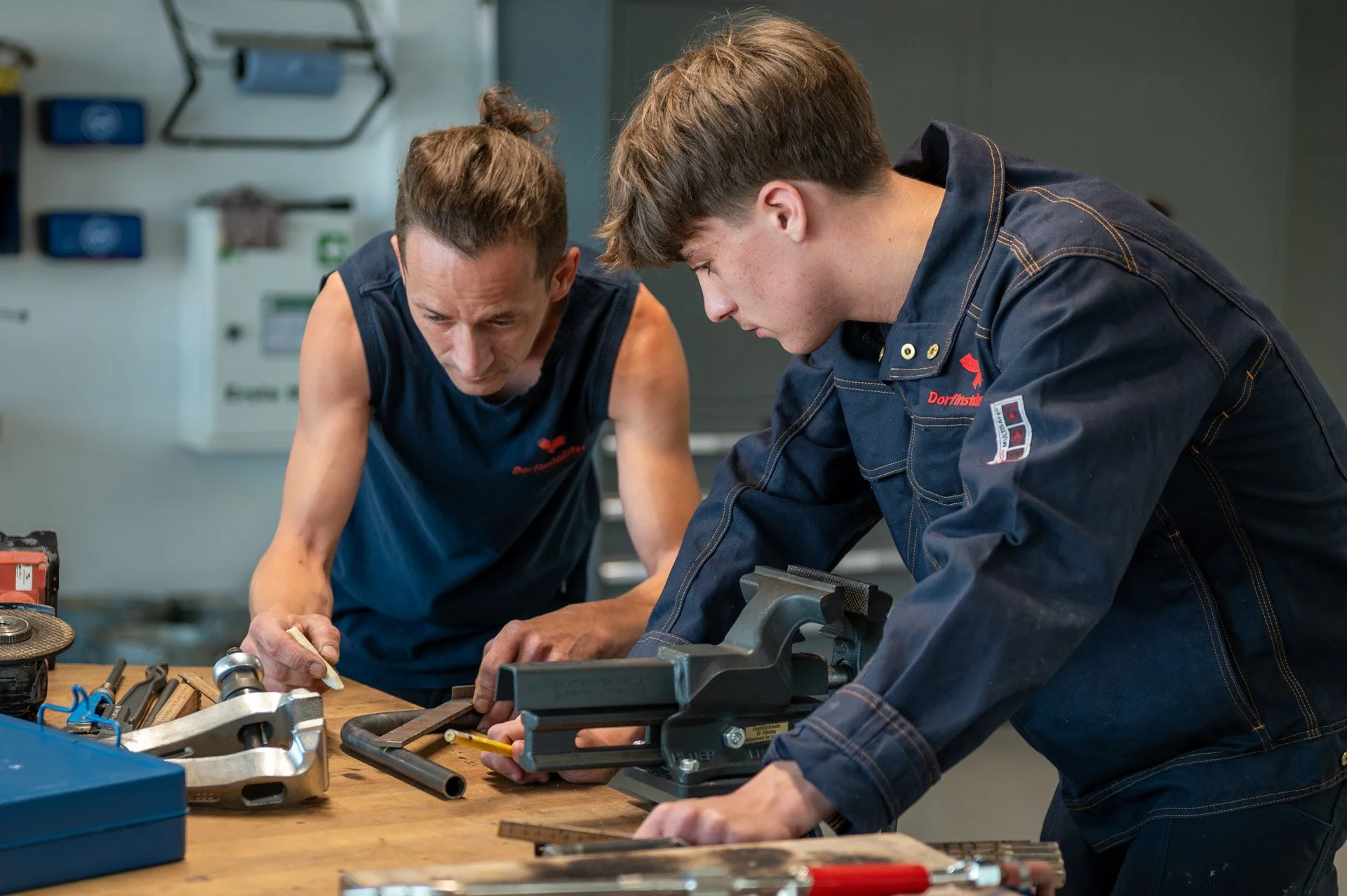 Zwei Männer arbeiten in einer Werkstatt an einer Arbeitstheke mit Werkzeugen und maschinellen Teilen.