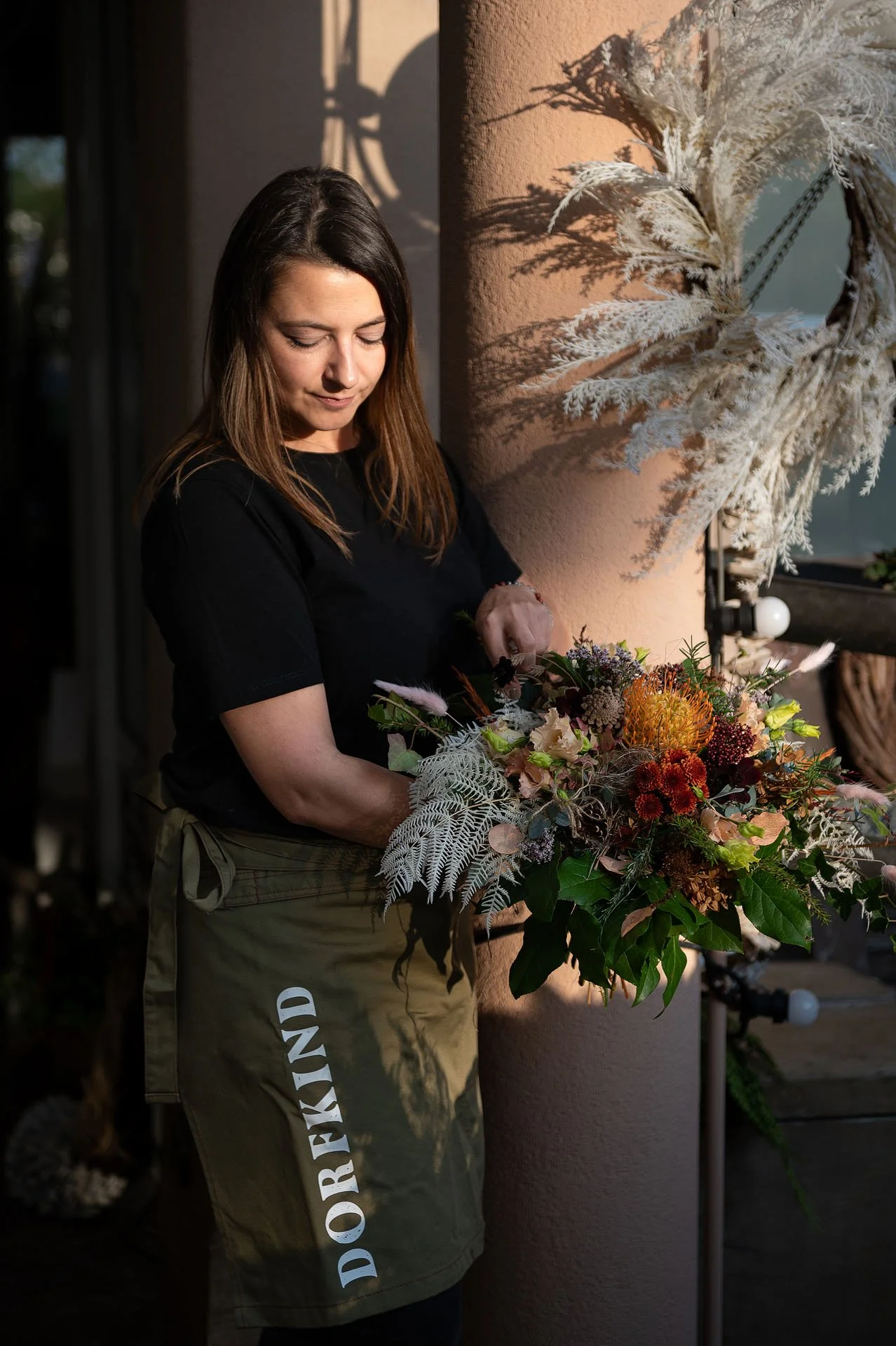 Eine junge Frau mit braunen Haaren trägt eine schwarze T-Shirt und eine grüne Hose, während sie einen bunten Blumenstrauß hält.