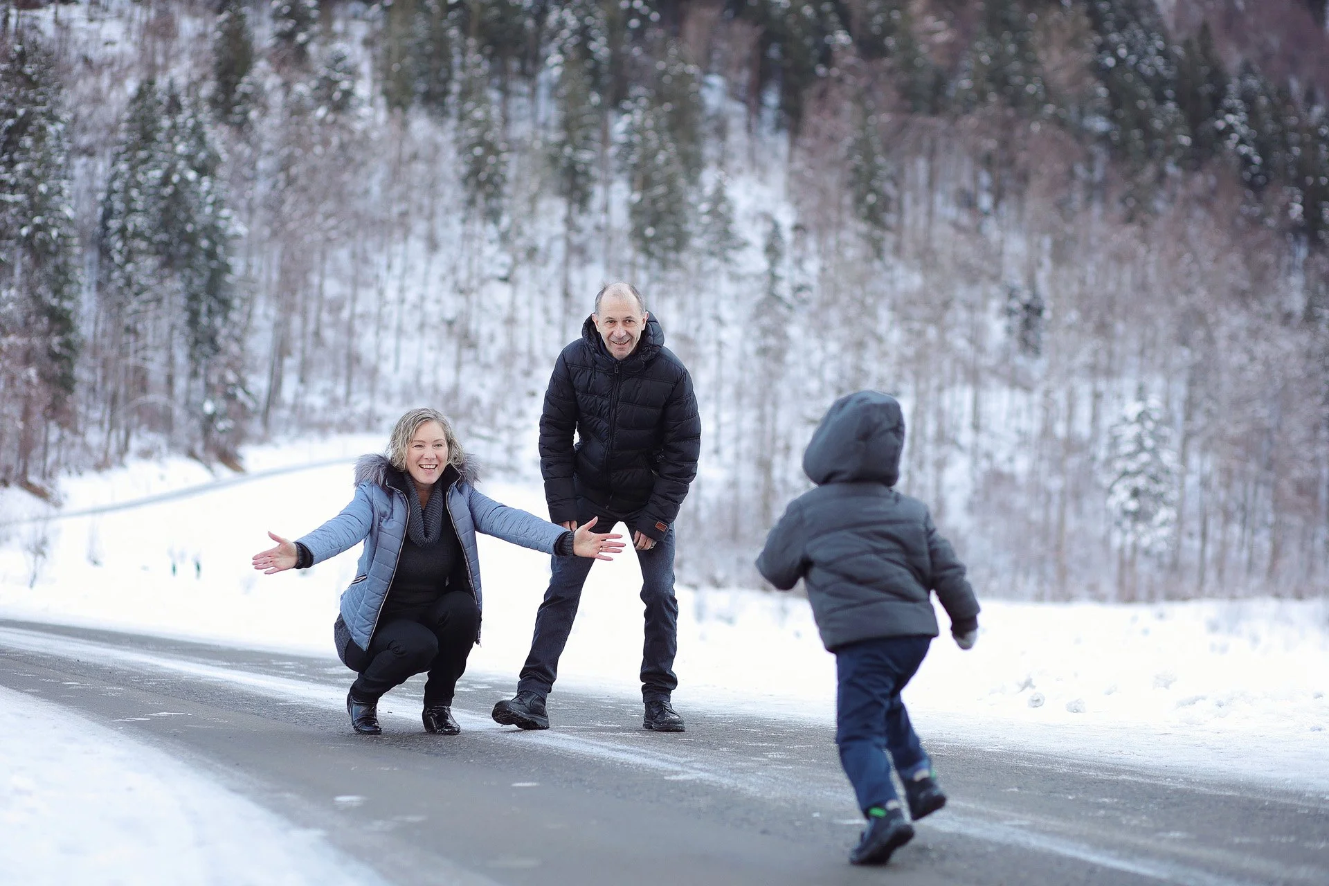 Familie beim Winterspaziergang im Schnee, die Frau ist glücklich und zieht Kinder am Spiel im Schnee, umgeben von verschneiten Bäumen.