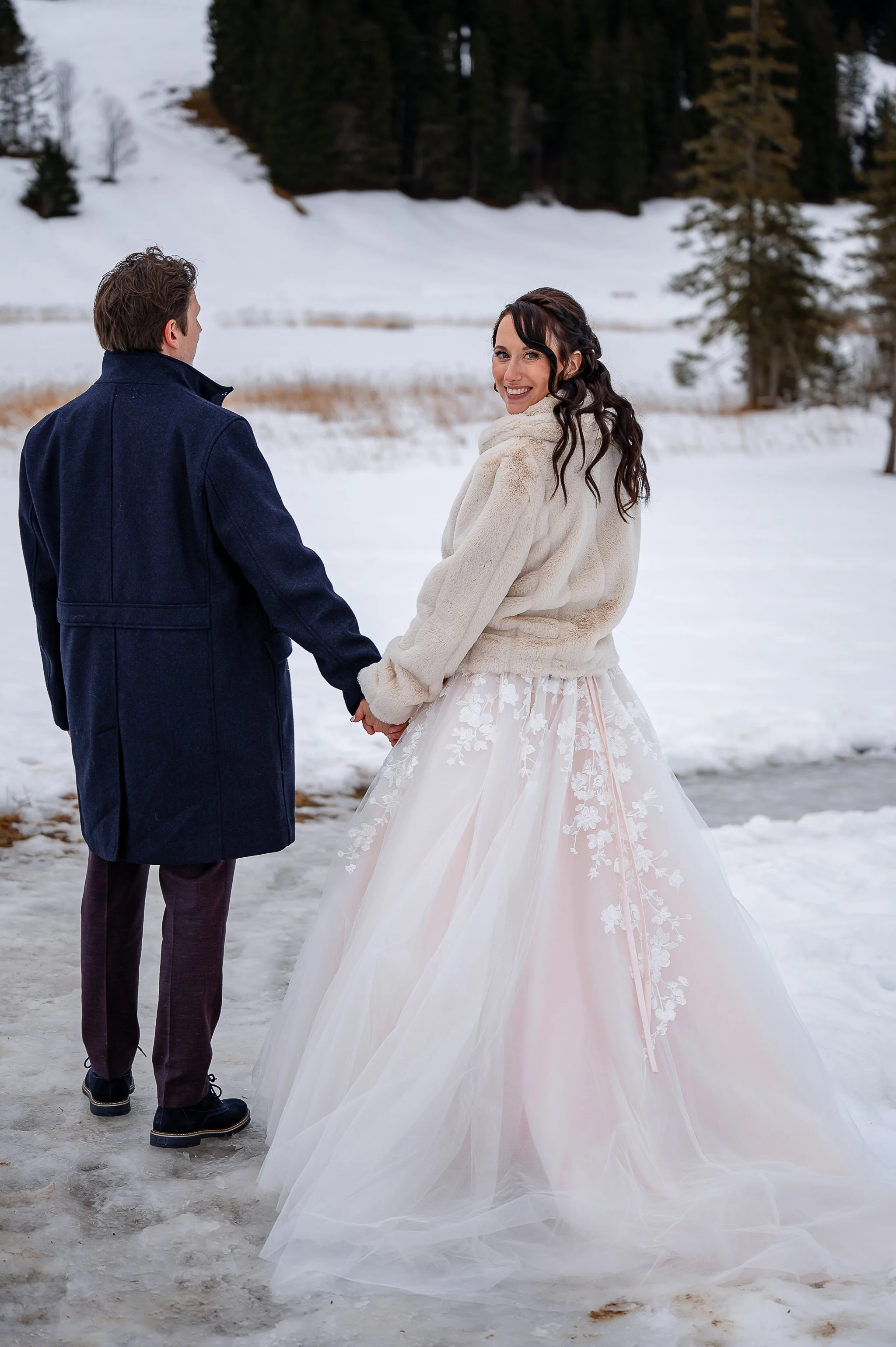 Ein Brautpaar hält sich an einem verschneiten Ort an den Händen, die Frau trägt ein Brautkleid mit Tüll und Stickerei, der Mann trägt einen dunklen Mantel.