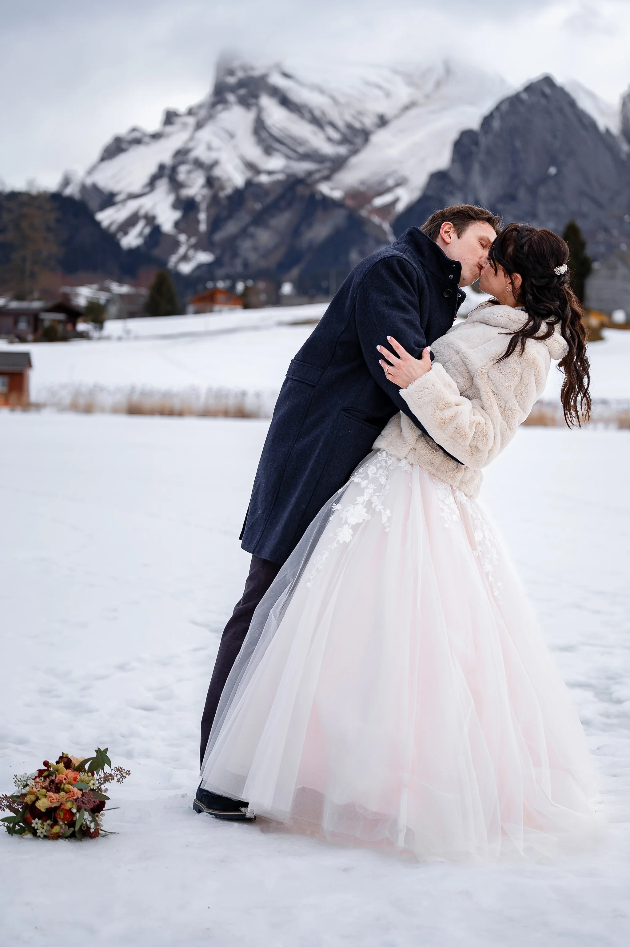 Ein Paar küsst sich in winterlicher Landschaft, die Frau trägt ein Hochzeitskleid, der Mann einen dunklen Mantel, im Hintergrund sind verschneite Berge.