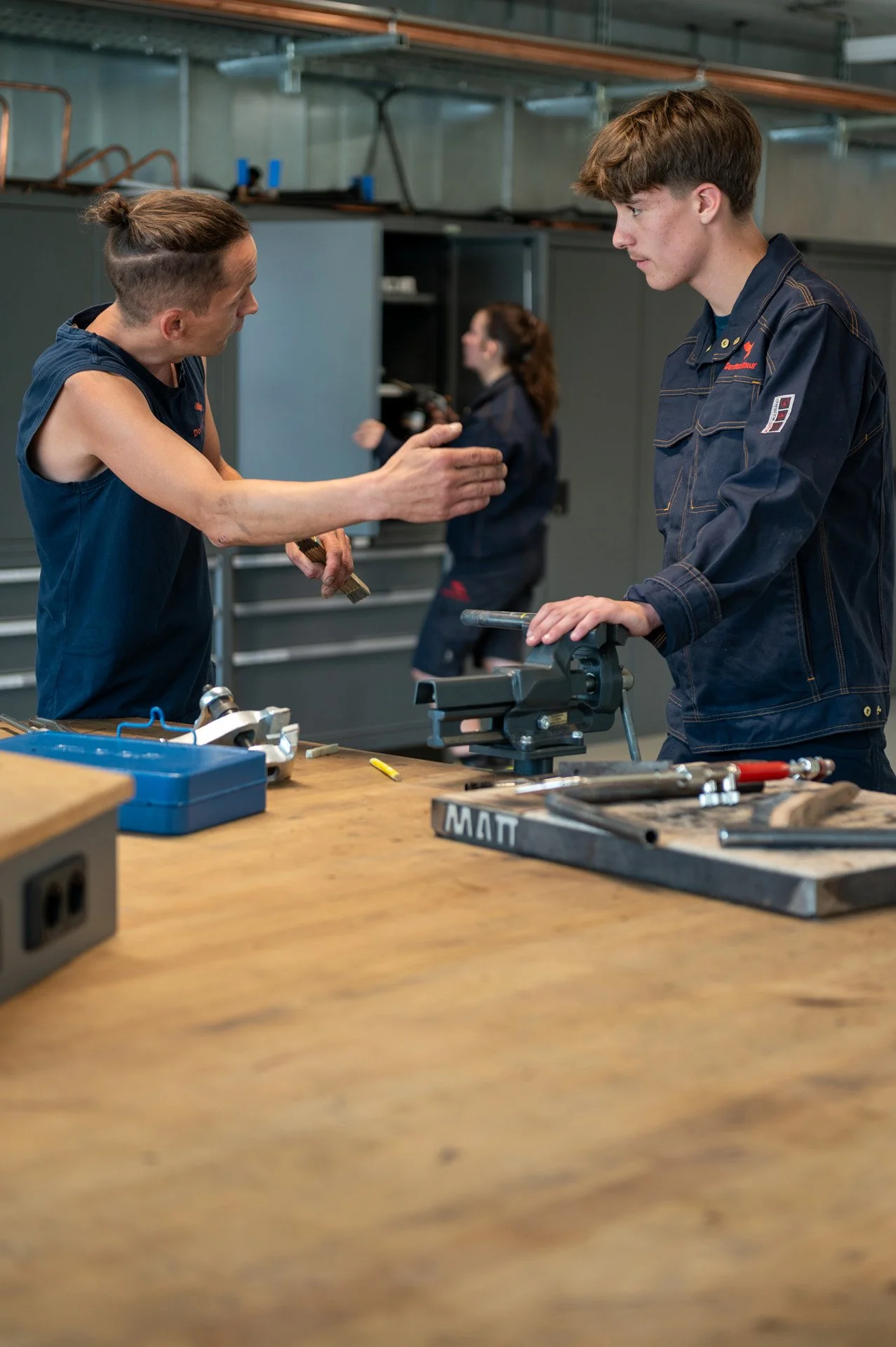 Zwei junge Menschen arbeiten in einer Werkstatt mit Werkzeugen, während eine Frau im Hintergrund ebenfalls arbeitet. Es gibt Werkbänke und Werkzeuge auf dem Tisch.