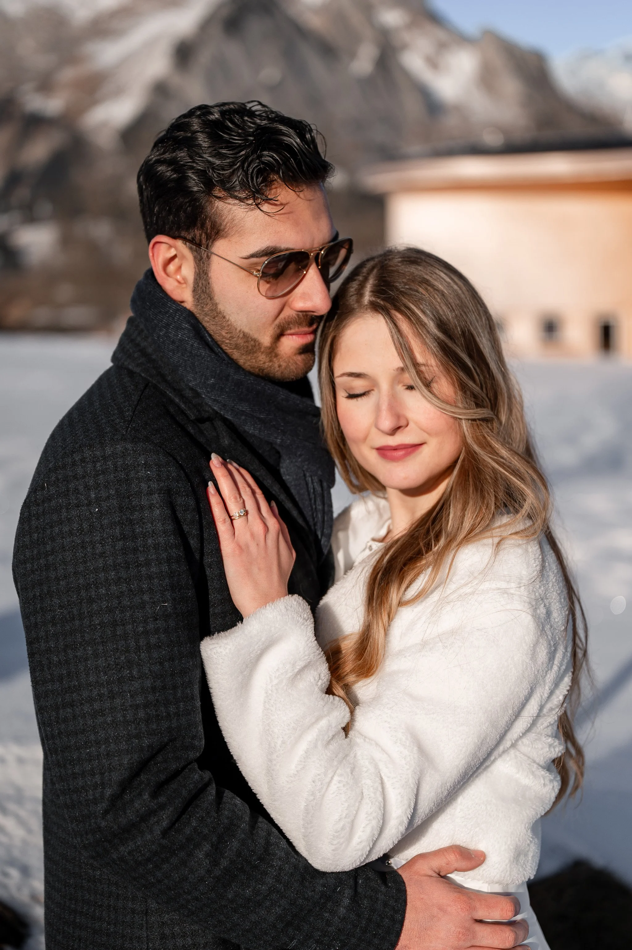 Ein Brautpaar umarmt sich im Schnee mit Bergen im Hintergrund, beide tragen Winterkleidung, die Frau hat geschlossene Augen, der Mann trägt Sonnenbrille.