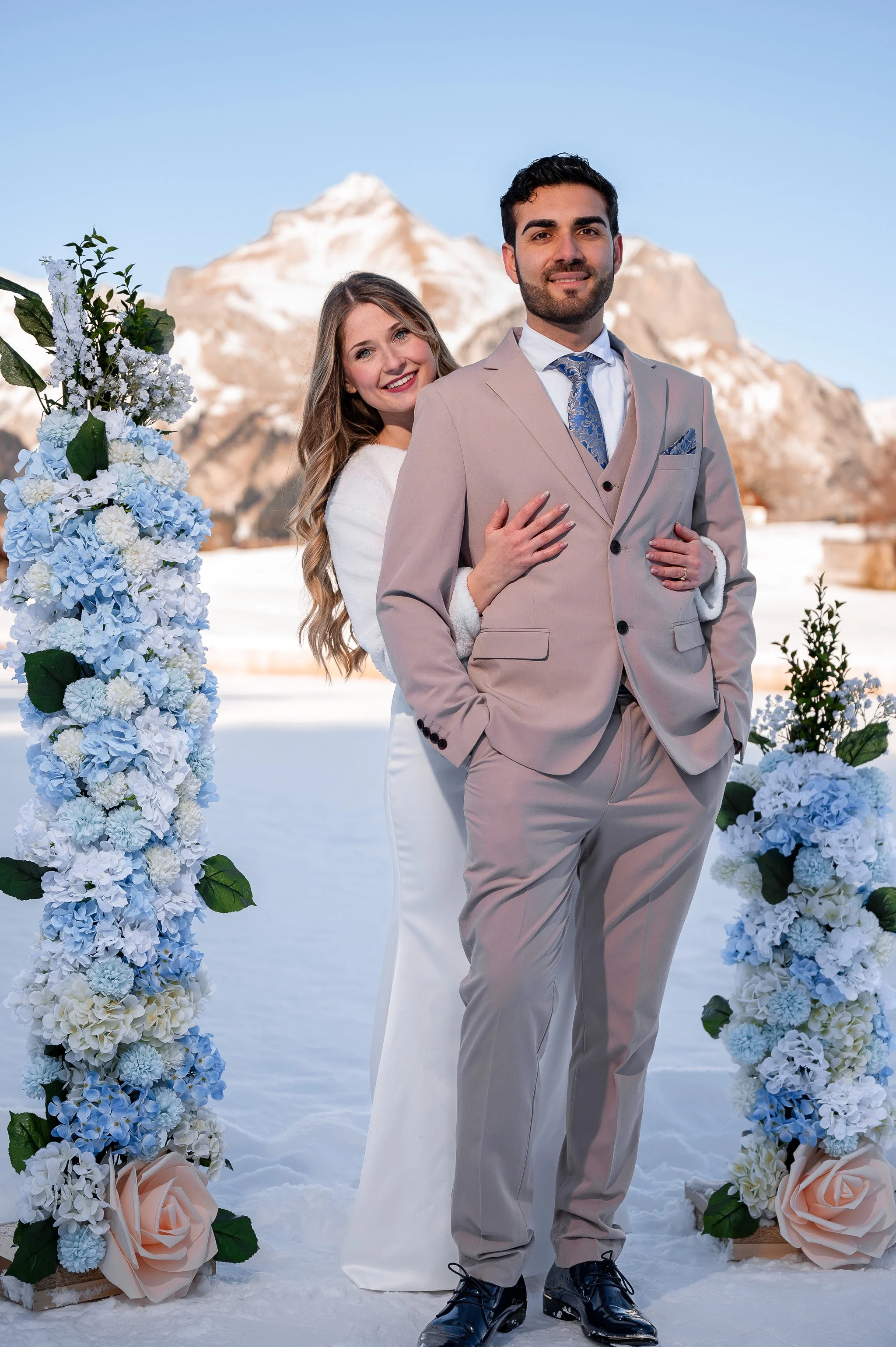 Ein Brautpaar in formeller Kleidung bei einer Winterhochzeit vor schneebedeckten Bergen, umgeben von Blumenarrangements.