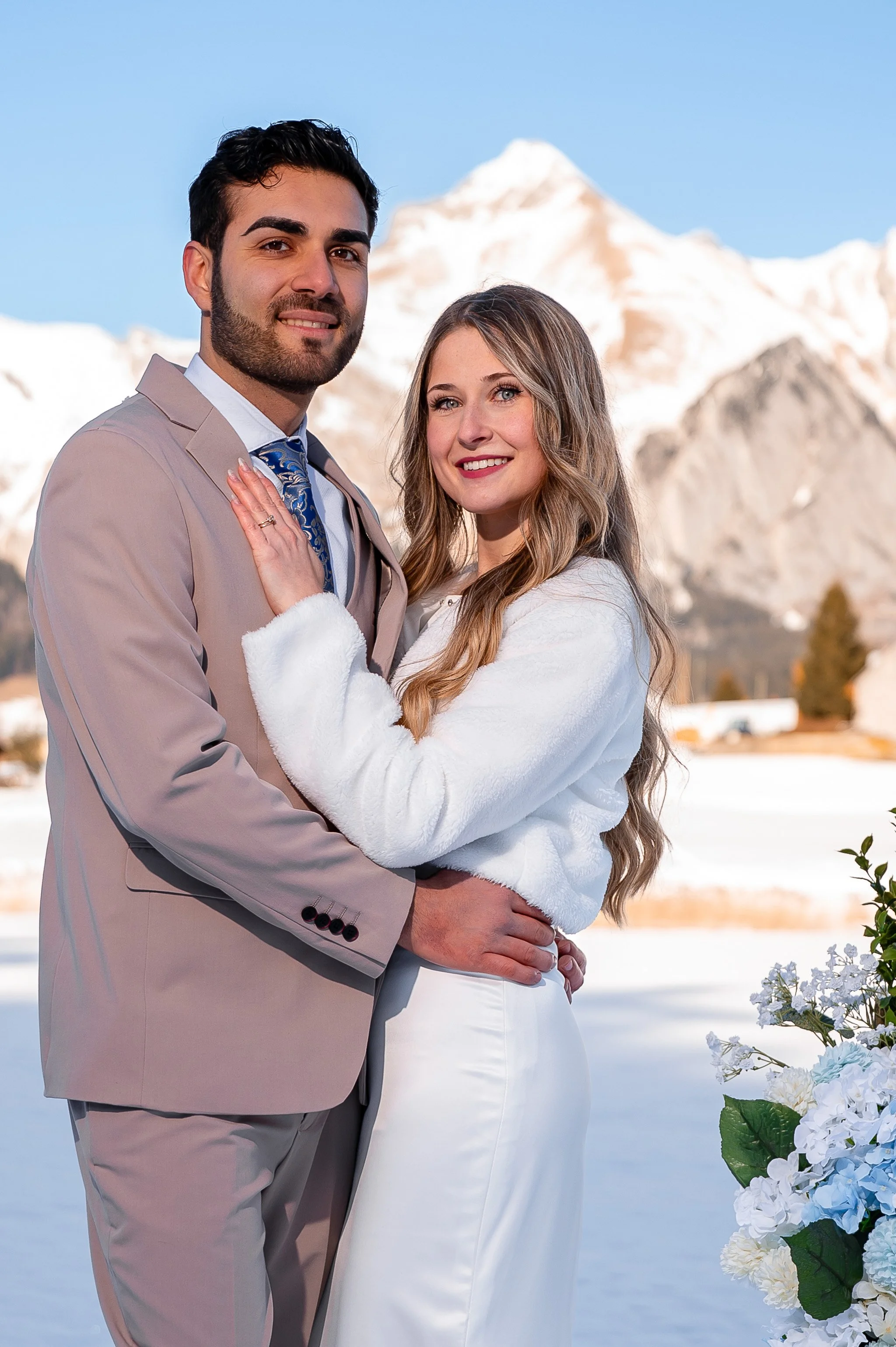 Ein Brautpaar steht in der Winterlandschaft vor schneebedeckten Bergen, umarmt sich und lächelt in die Kamera.