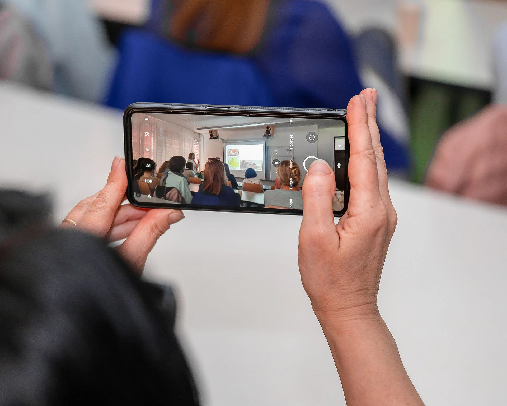Nahaufnahme einer Person, die ein Smartphone benutzt, um ein Bild eines Classroom-Settings zu machen, mit mehreren Schülern und einem Lehrer, der auf eine Präsentation zeigt.