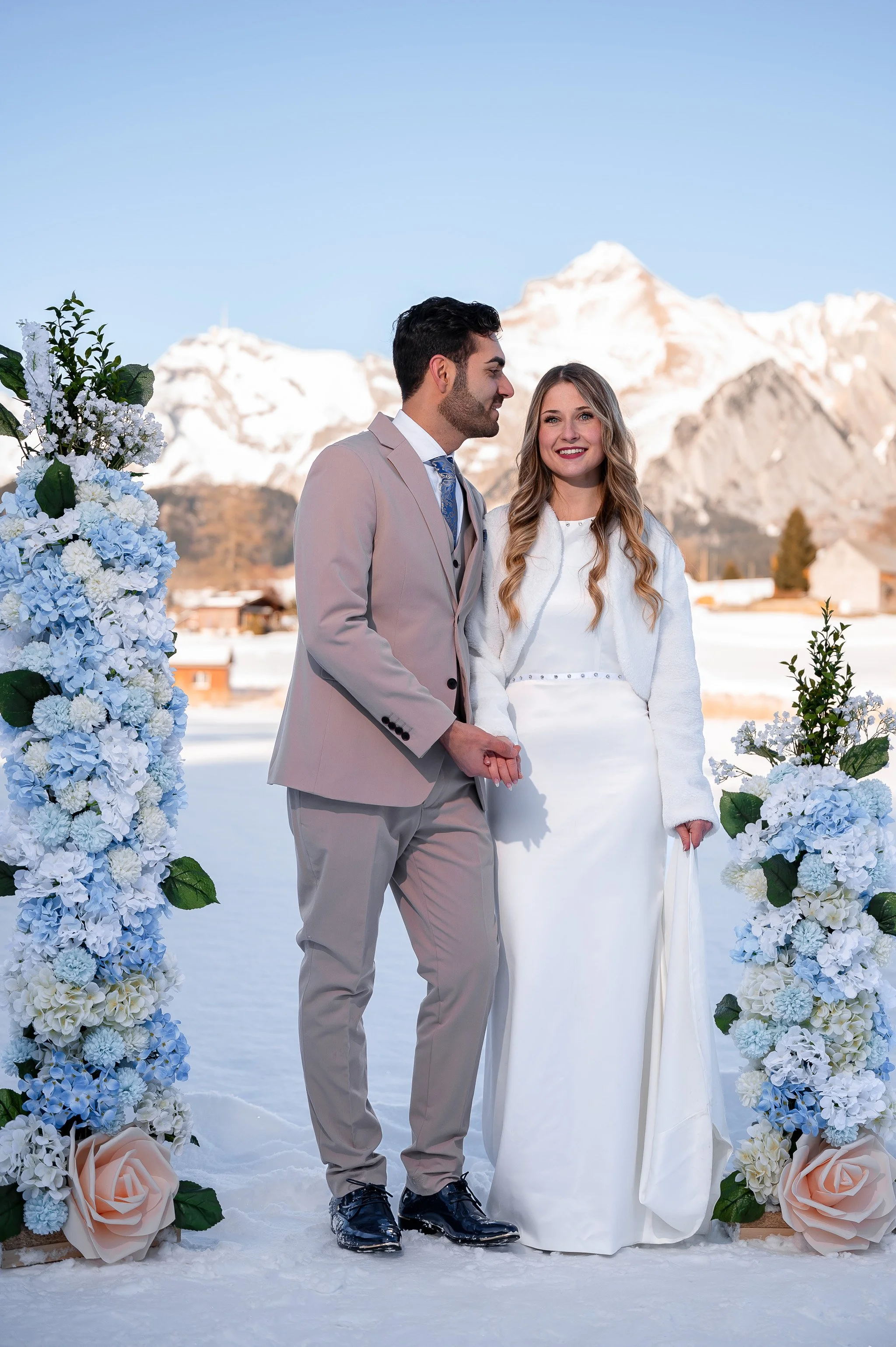 Ein glückliches Brautpaar bei einer Winterhochzeit unter einem blauen Himmel mit schneebedeckten Bergen im Hintergrund, umgeben von blauen und weißen Blumen