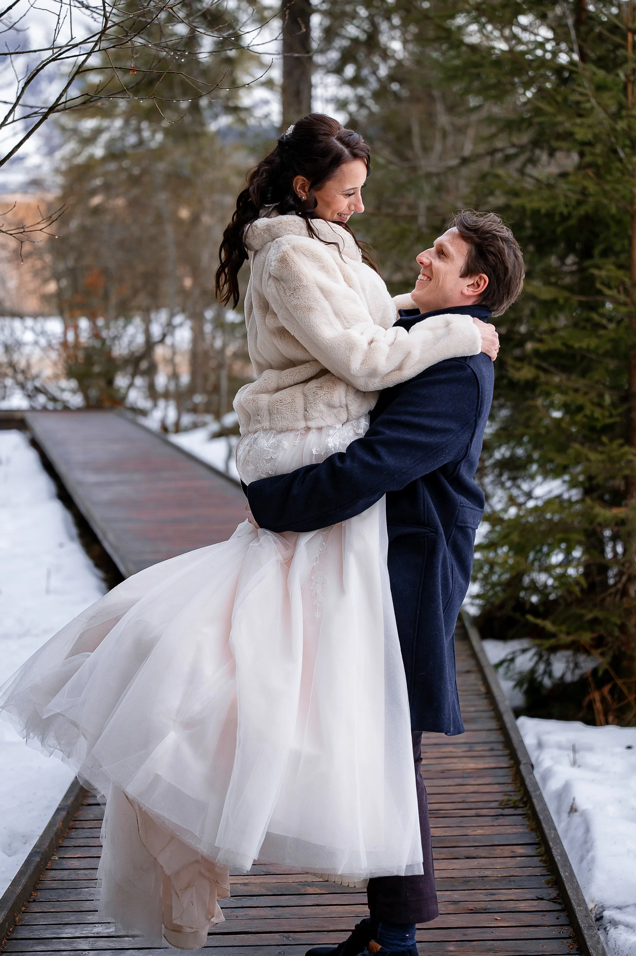 Ein Brautpaar in winterlicher Landschaft, das sich liebevoll ansieht. Der Bräutigam hebt die Braut auf, die in einem weißen Brautkleid und einem weißen Pelzmantel gekleidet ist.