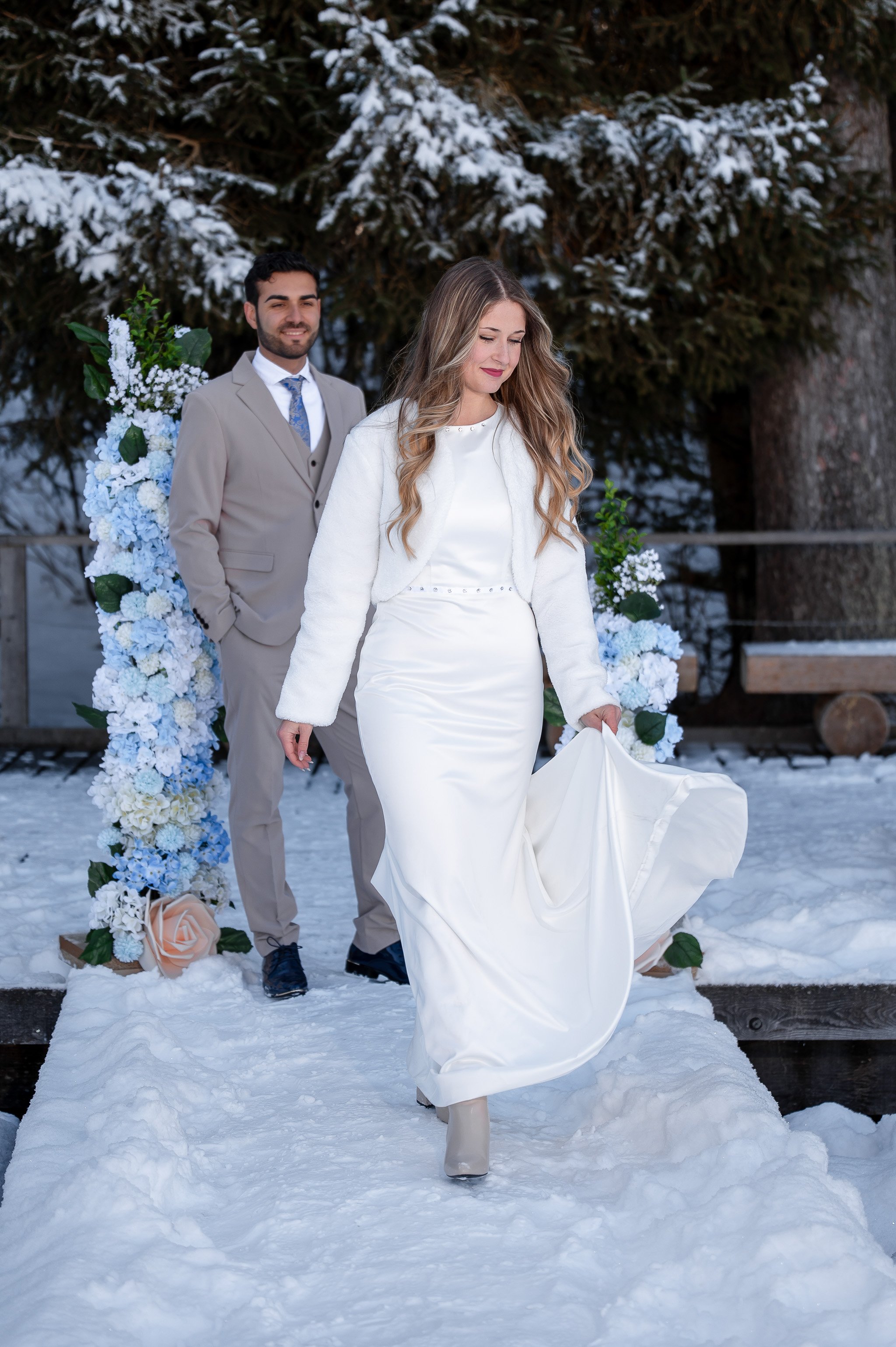 Braut in weißem Kleid und Jacke, Brautpaar im Schnee bei Hochzeit, hinter Blumenbogen mit weißen und blauen Blumen, im Winter im Freien.