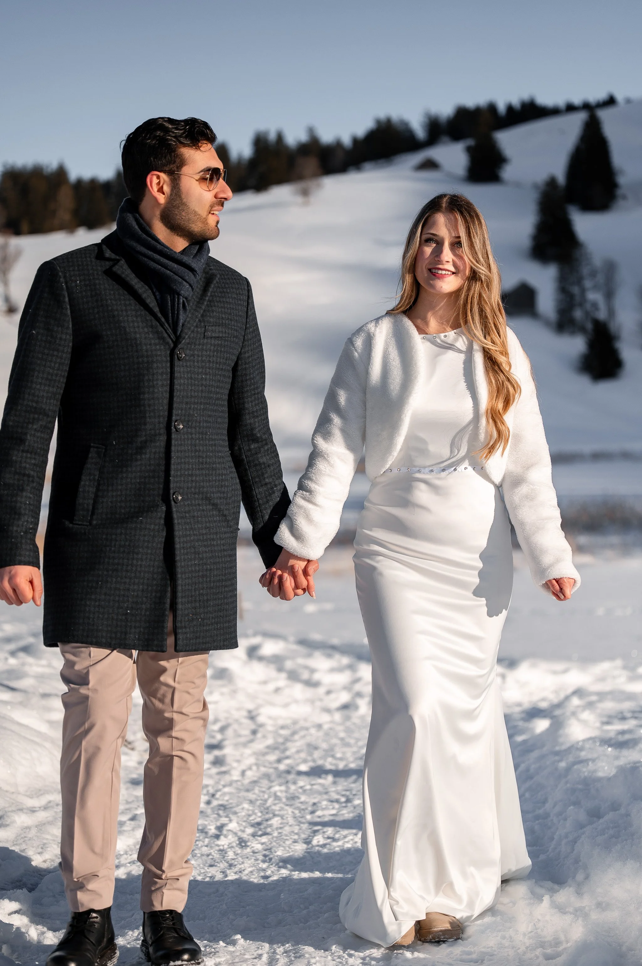 Ein Brautpaar hält Händchen im Schnee in einer winterlichen Landschaft, sie trägt ein weißes Hochzeitskleid und er einen dunklen Mantel mit Sonnenbrille.