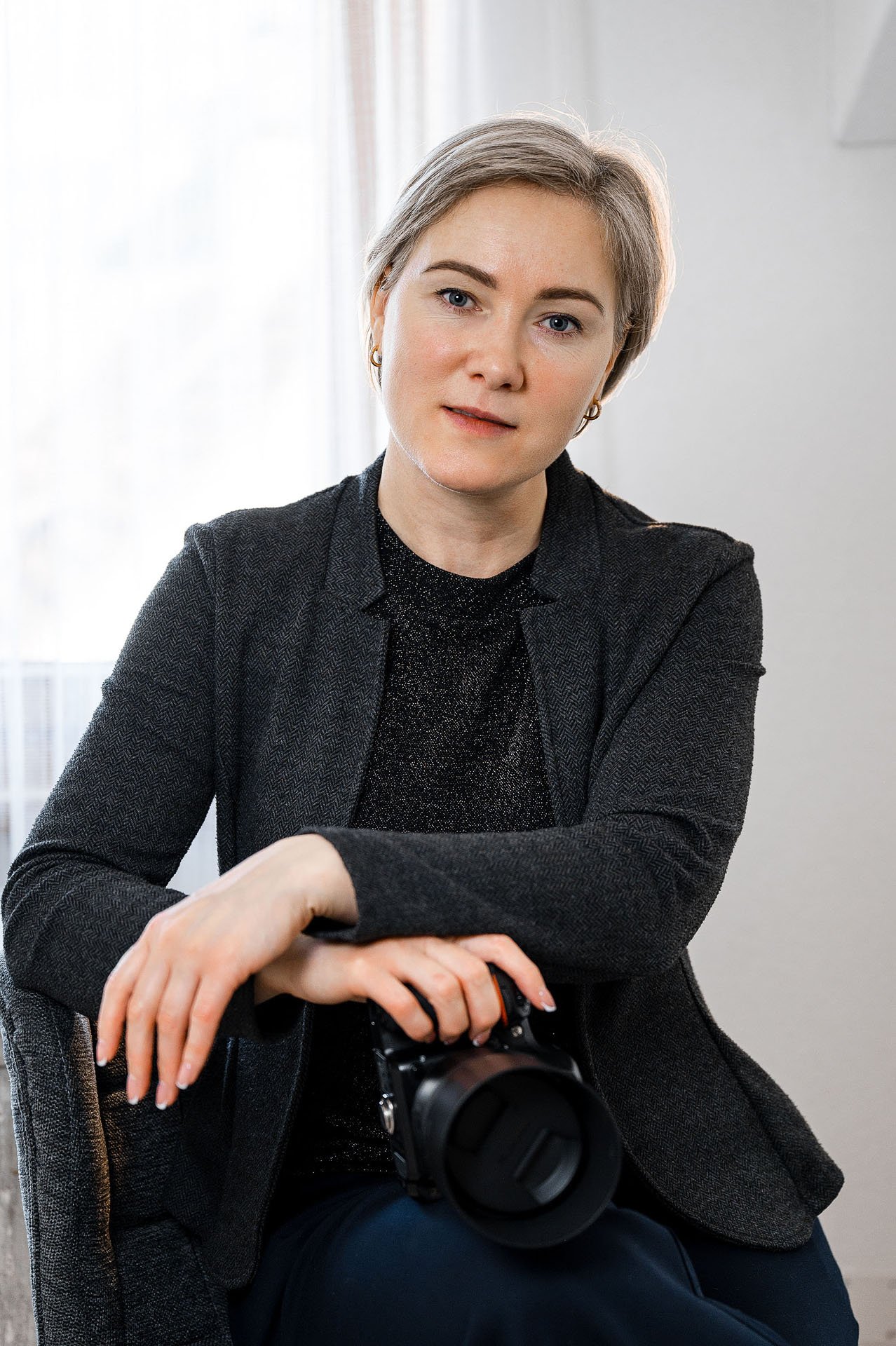 Eine Frau mit kurzen, grauen Haaren und blauen Augen, die in einem dunklen Blazer sitzt und eine Kamera in den Händen hält.