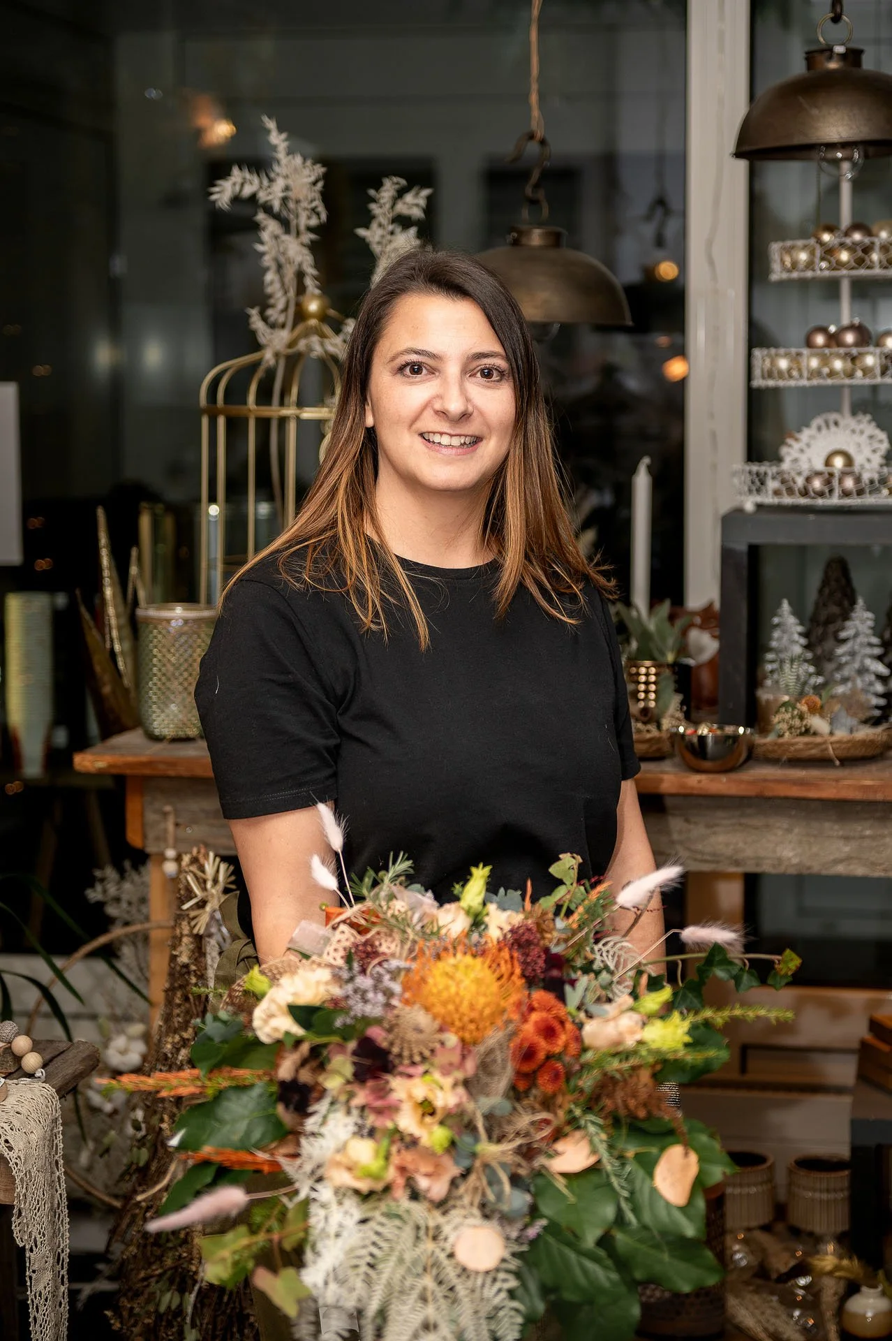 Eine lachende Frau mit braunen Haaren in einem schwarzen T-Shirt steht hinter einem bunten Blumenstrauß in einem dekorativen Geschäft mit dunklem, gemütlichem Ambiente.