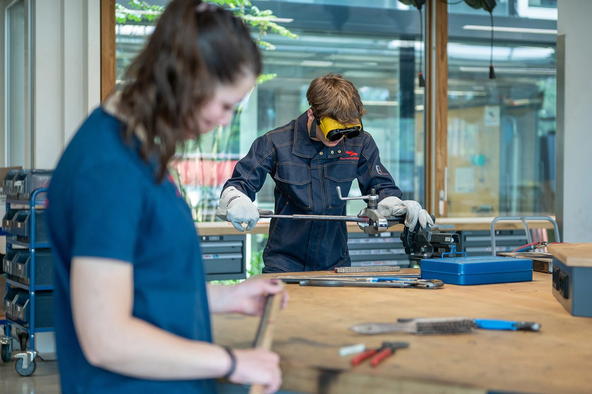 Zwei Menschen arbeiten in einer Werkstatt, einer benutzt eine Schutzbrille und arbeitet mit einem Werkzeug, der andere beobachtet. Es gibt Werkbänke und Werkzeug in der Umgebung.