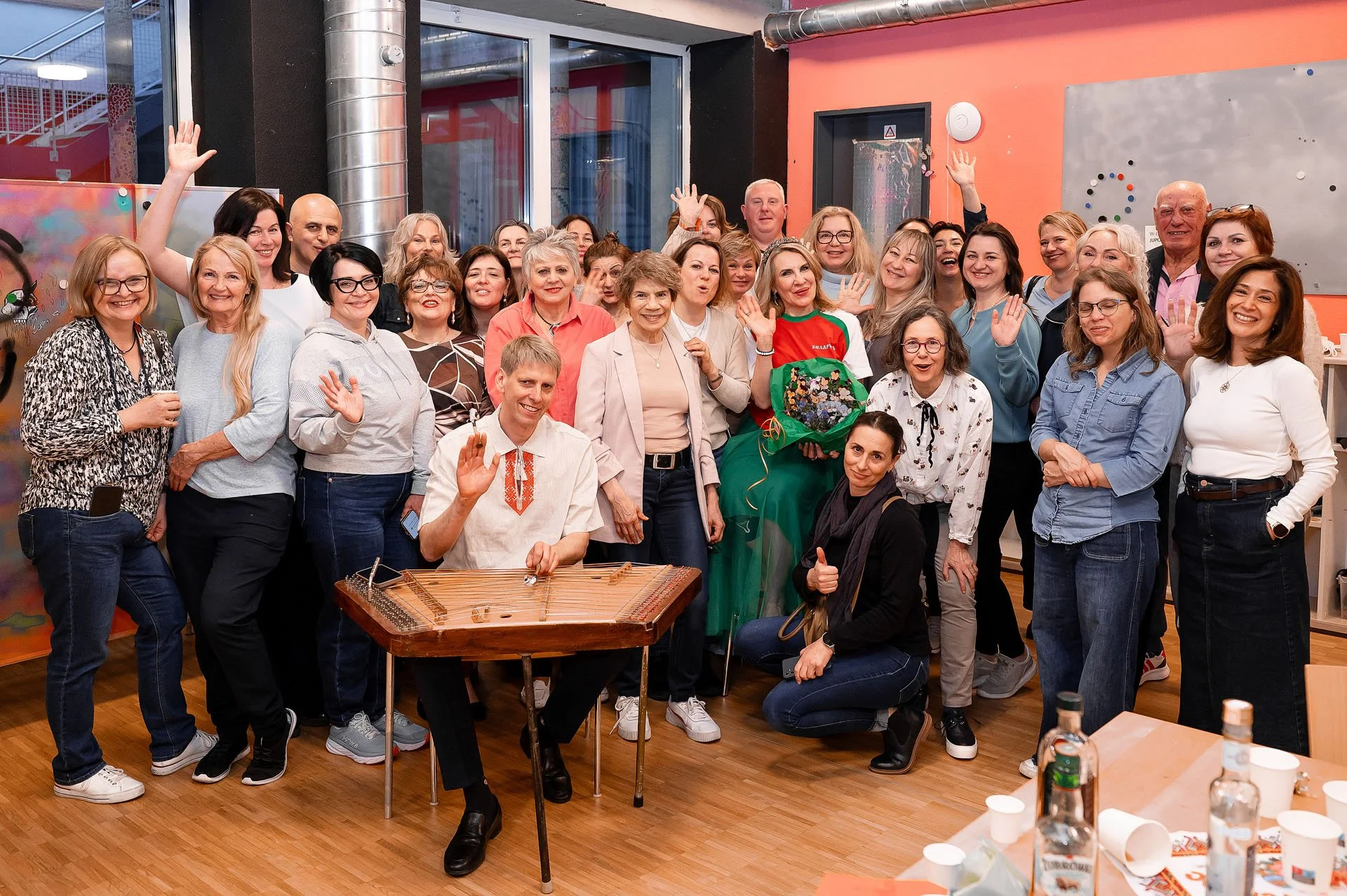 Gruppe von Menschen in einem Raum, einige winken und lächeln, im Hintergrund bunte Wände und Whiteboards, im Vordergrund ein Tisch mit Wasserflaschen und Papptellern.