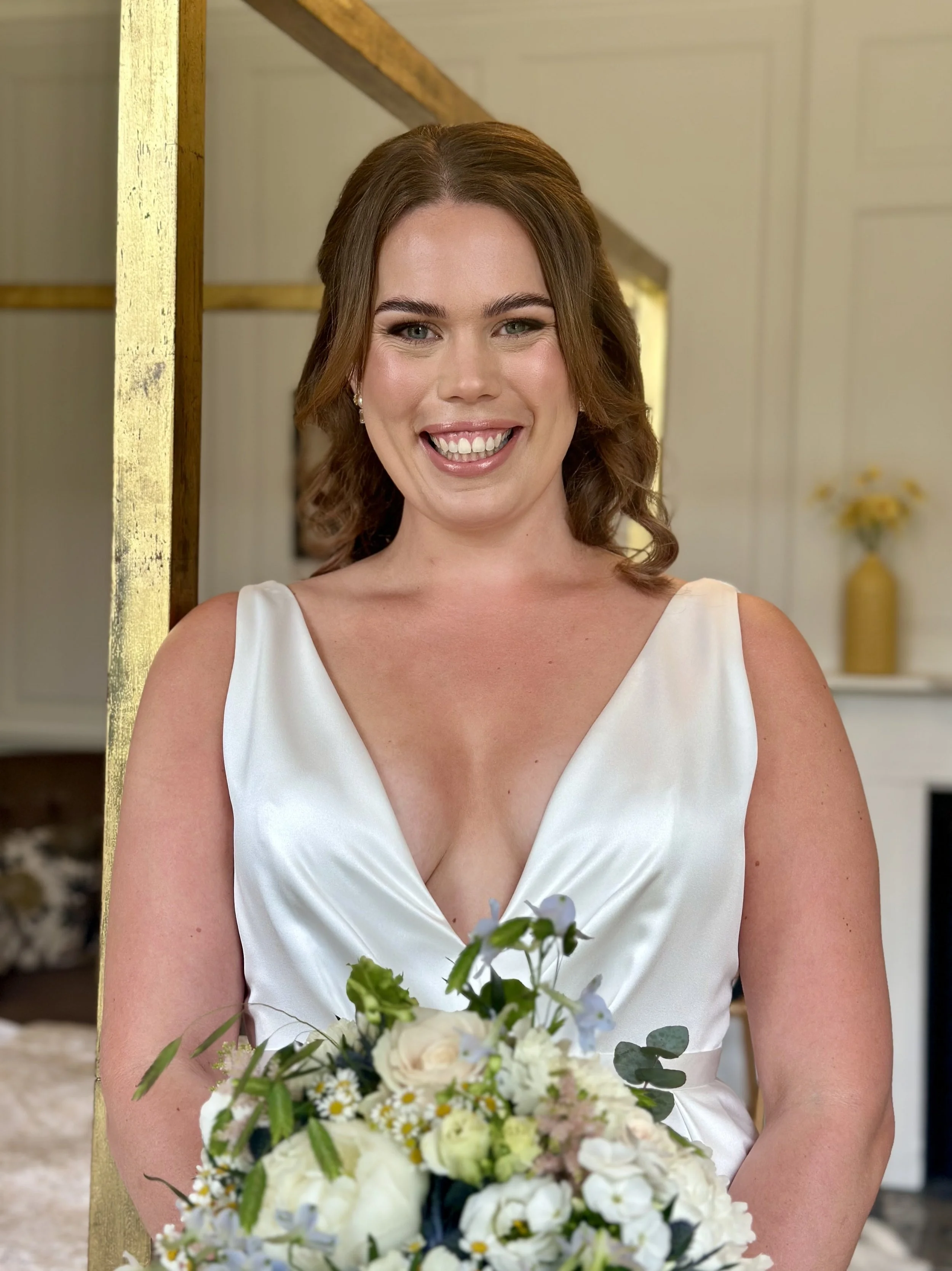 A woman in a white wedding dress holding a bouquet of white and light flowers, smiling in a well-lit indoor setting.