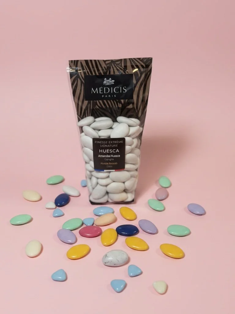 Une pochette de bonbons colorés et des bonbons blancs en forme d'amandes sur un fond rose.