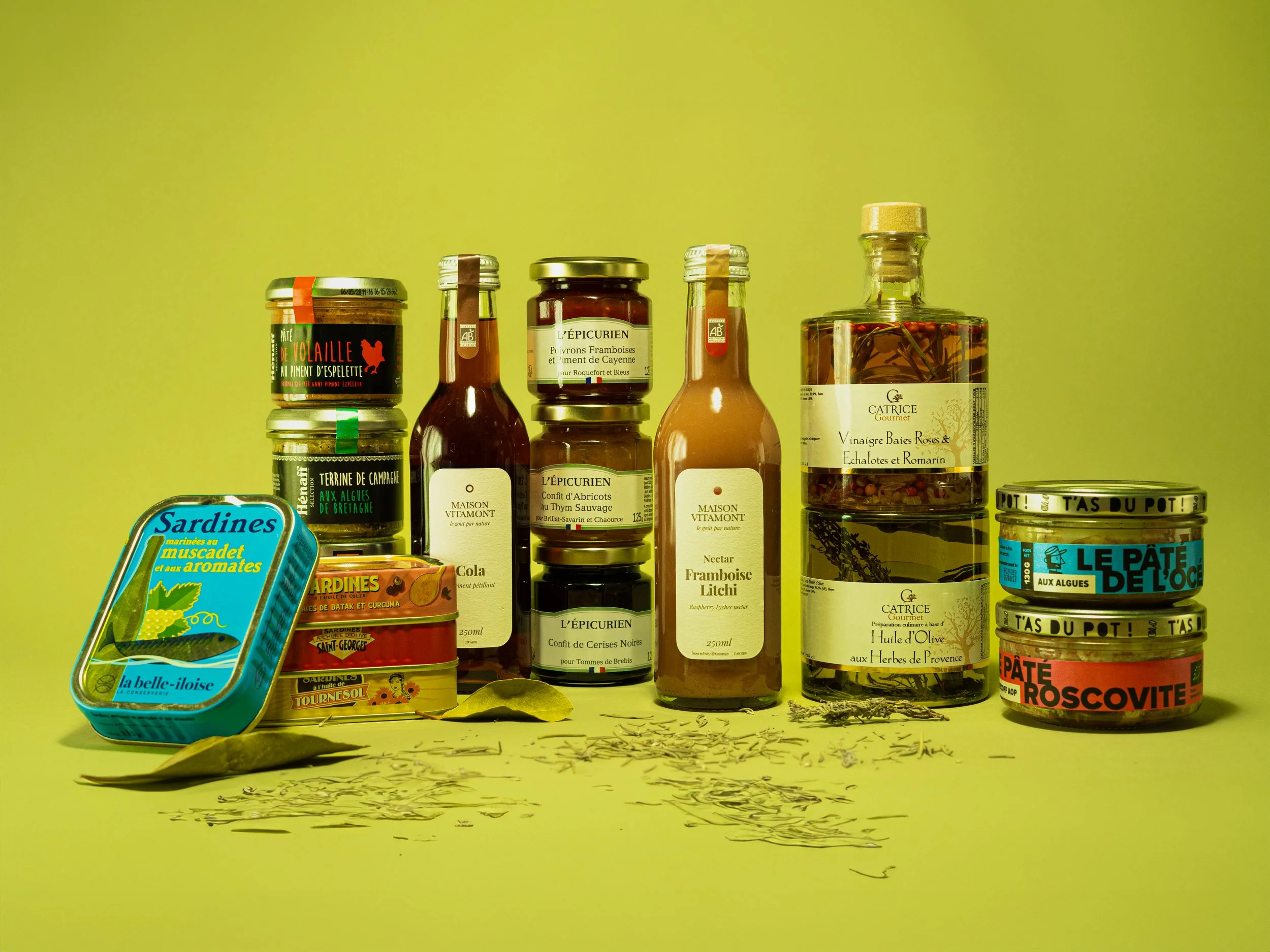 Une collection de conserves, bocaux et fioles alimentaires, comprenant des sardines, des pâtés, des sauces, des confitures, et des épices, disposés sur un fond jaune.